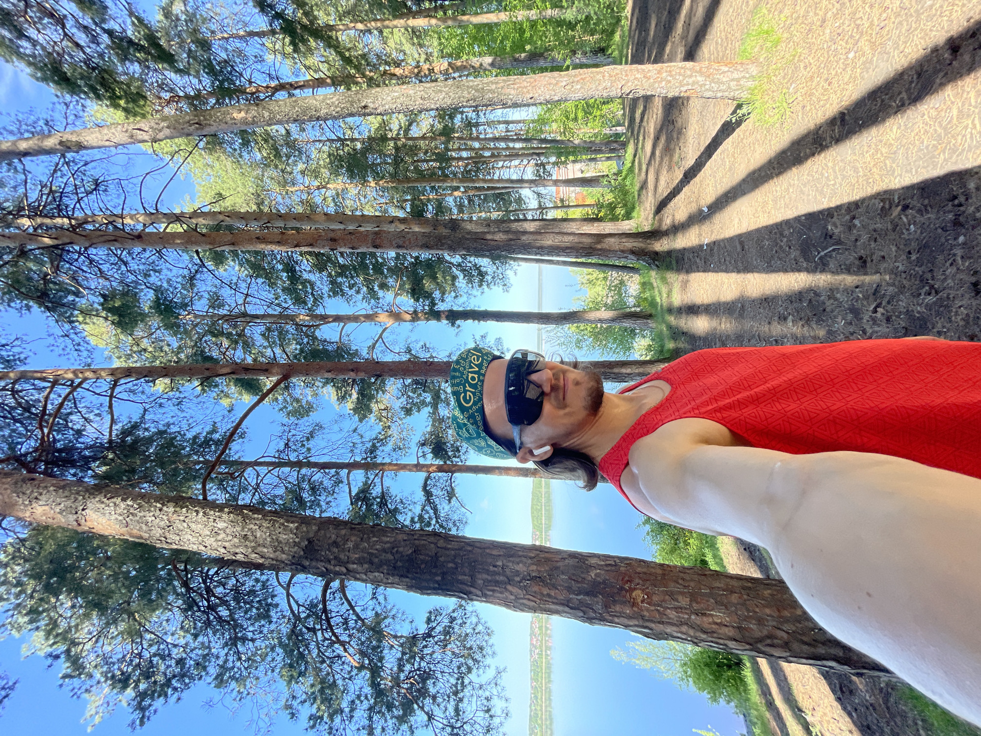 The image is a selfie taken outdoors, presumably in a park setting. The man in the foreground is wearing a red tank top, sunglasses, a baseball cap, and appears to have an earbud in his ear. He is positioned next to a large tree trunk. The background shows a row of tall pine trees against a clear blue sky. The water can be seen in the background, likely a part of the Izhevsk reservoir, which defines the shoreline as the scene is in the city of Izhevsk, Russia. Sunlight is creating long shadows across the ground. The angle of the photo suggests it was taken at a low angle, looking upwards towards the trees and sky.