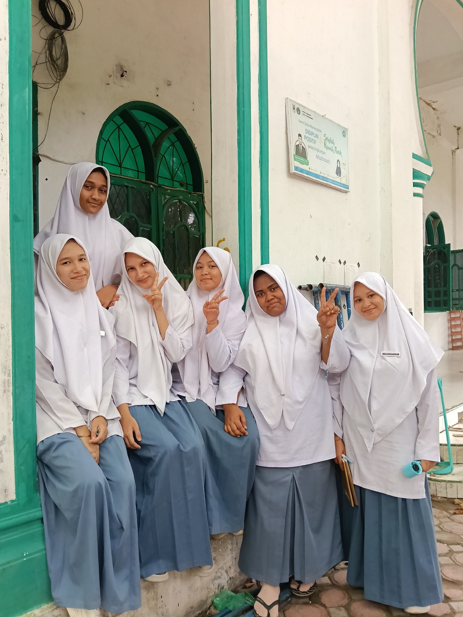 A group of six female students wearing white hijabs and long blue skirts pose together on a step in Lhokseumawe, Indonesia. The location appears to be a building with green trim around windows and doors. A sign written in Indonesian is visible on the wall. Some of the students are flashing the peace sign.