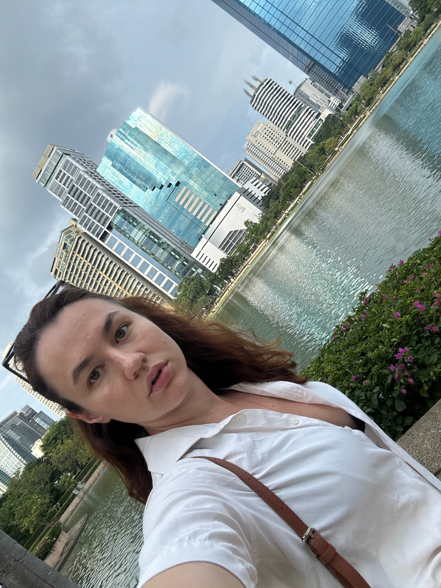 The image is a selfie taken by a woman with brown hair, wearing a white button-up shirt and a brown crossbody bag. She is looking at the camera with a neutral expression. The background shows a body of water, lush green trees, and several modern high-rise buildings. The overall scene suggests an urban park setting within Bangkok, Thailand, possibly Lumpini Park, given the characteristic combination of a large lake, abundant greenery, and prominent skyscrapers. The sky is overcast.