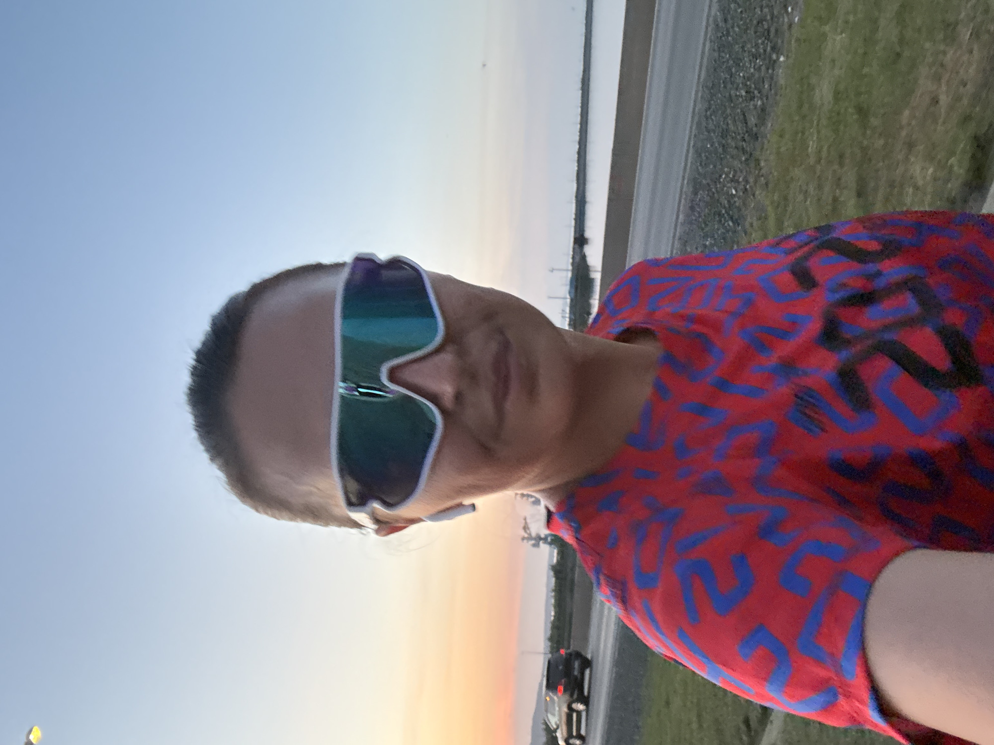 A person wearing sunglasses and a red shirt with blue geometric patterns is positioned outdoors. The person is likely standing near a road or highway with a guardrail visible in the background. A car is driving on the road. The sky is transitioning from daylight to dusk, displaying shades of orange and light blue. The location is likely Chaikovsky, Russia.