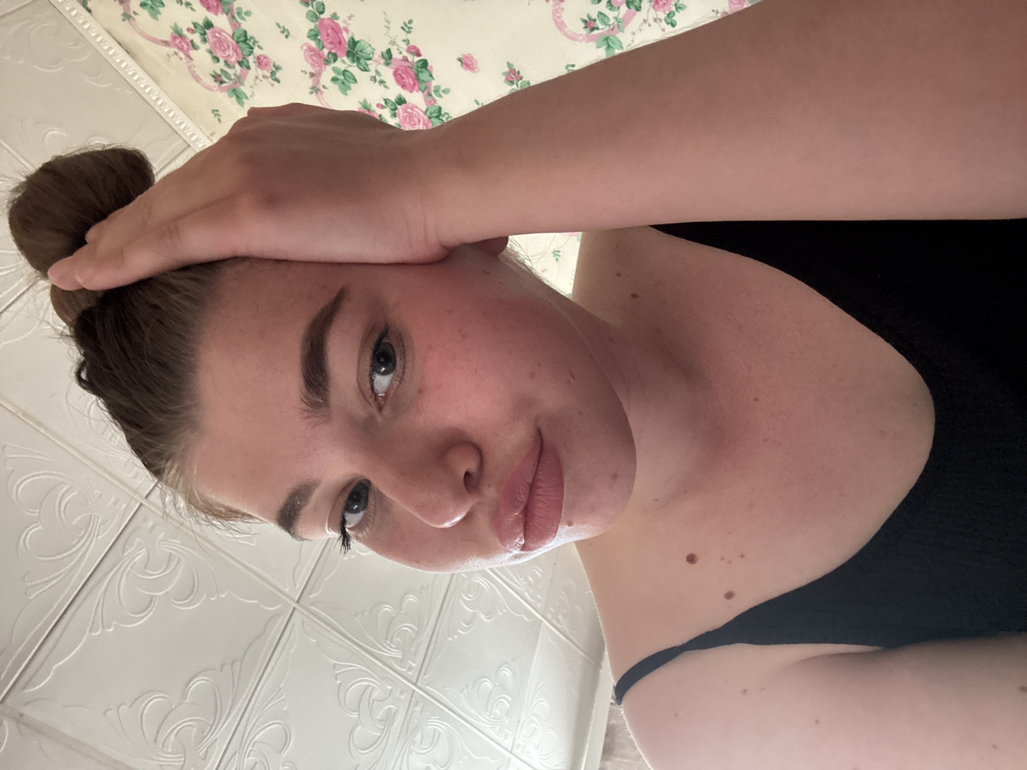 A young woman is posing for a selfie-style picture. She has brown hair in a bun and is wearing a black top. Her right hand is resting on her forehead. The background consists of a floral patterned surface and a white, textured wall. No location is evident.