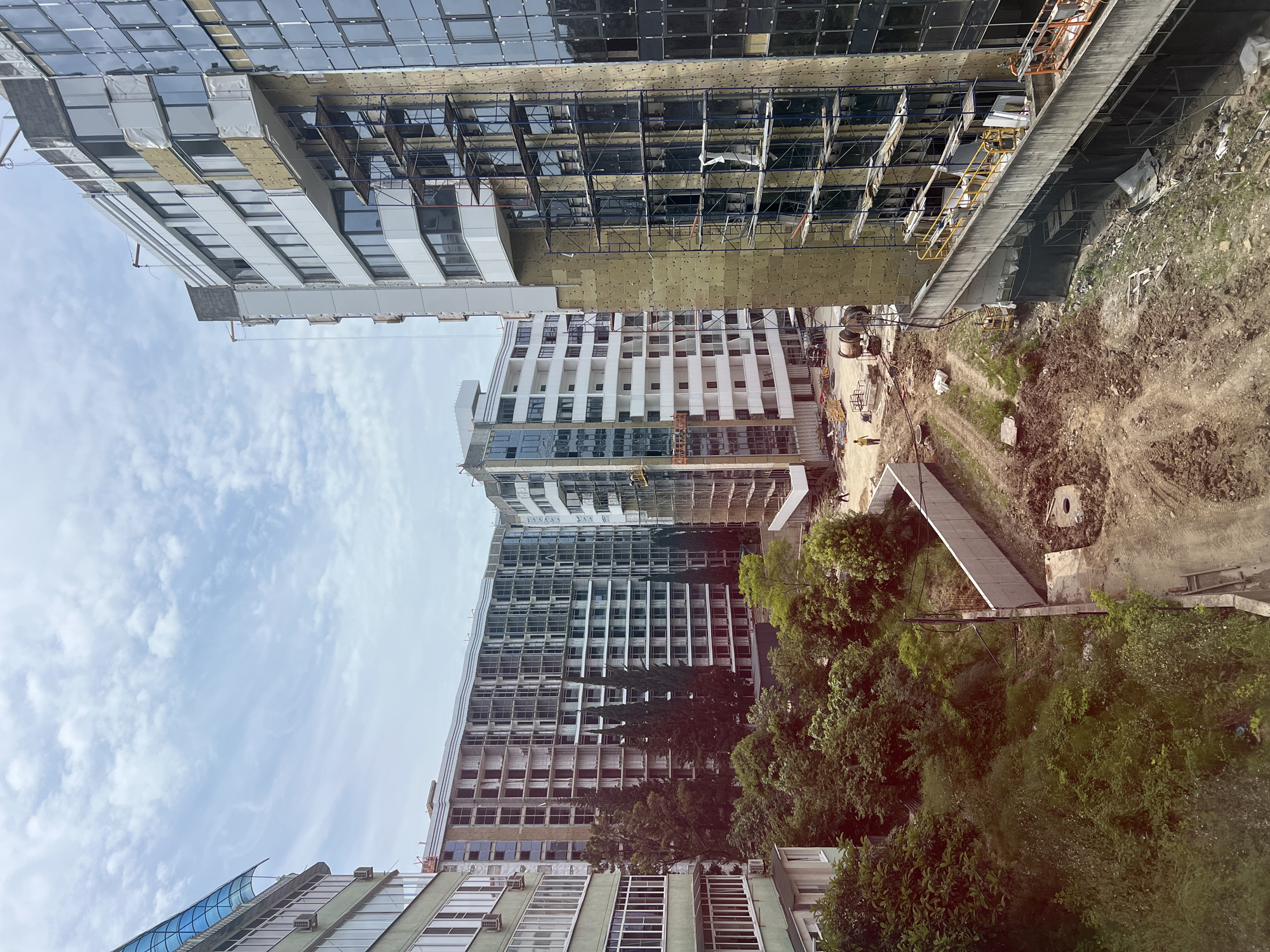 This is a high-angle, full shot of a construction site in Sochi, Russia, featuring several multi-story buildings under various stages of completion. The foreground shows a dirt path with some construction equipment and debris. A concrete retaining wall runs along the right side of the path, leading to a large building in the mid-ground that is covered in scaffolding and insulation. This building has a partially completed glass facade. To the left of this building, more residential buildings are visible, some appearing further along in their construction process with windows installed and balconies present. The background features a cloudy sky with patches of blue. Trees and greenery are interspersed with the buildings, suggesting a semi-urban or developing area. One person in a yellow shirt is visible walking on the dirt path.