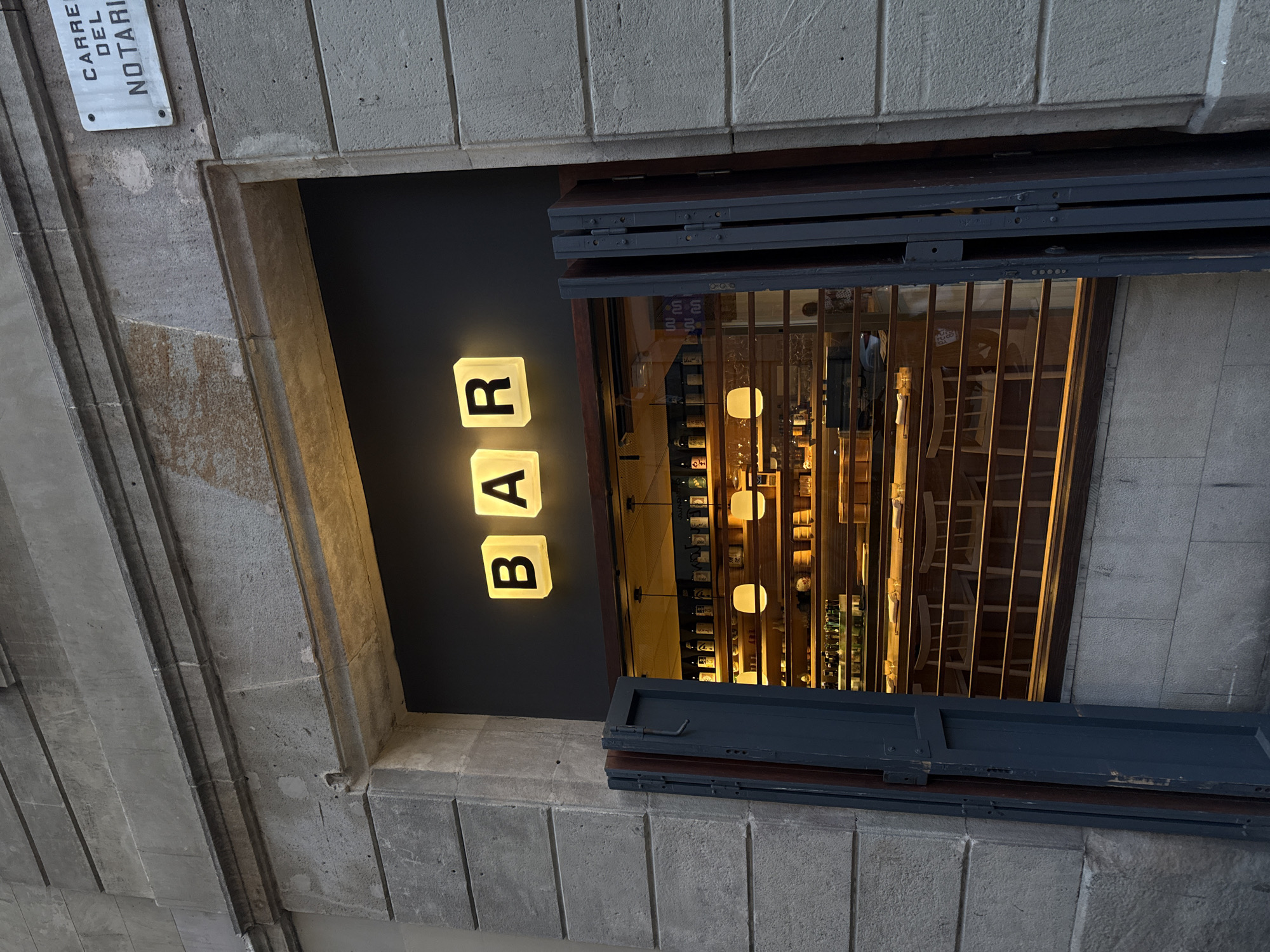 The image shows the exterior of a bar in Barcelona, Spain. A sign reading "BAR" is illuminated in block letters next to a window. The window is partially shuttered and provides a view of the bar's interior, including shelves stocked with bottles and hanging lights. A street sign that reads "CARRE DEL NOTARI" is visible on the building exterior above the window. The architecture is typical of the city, with stone facades and a narrow street layout.