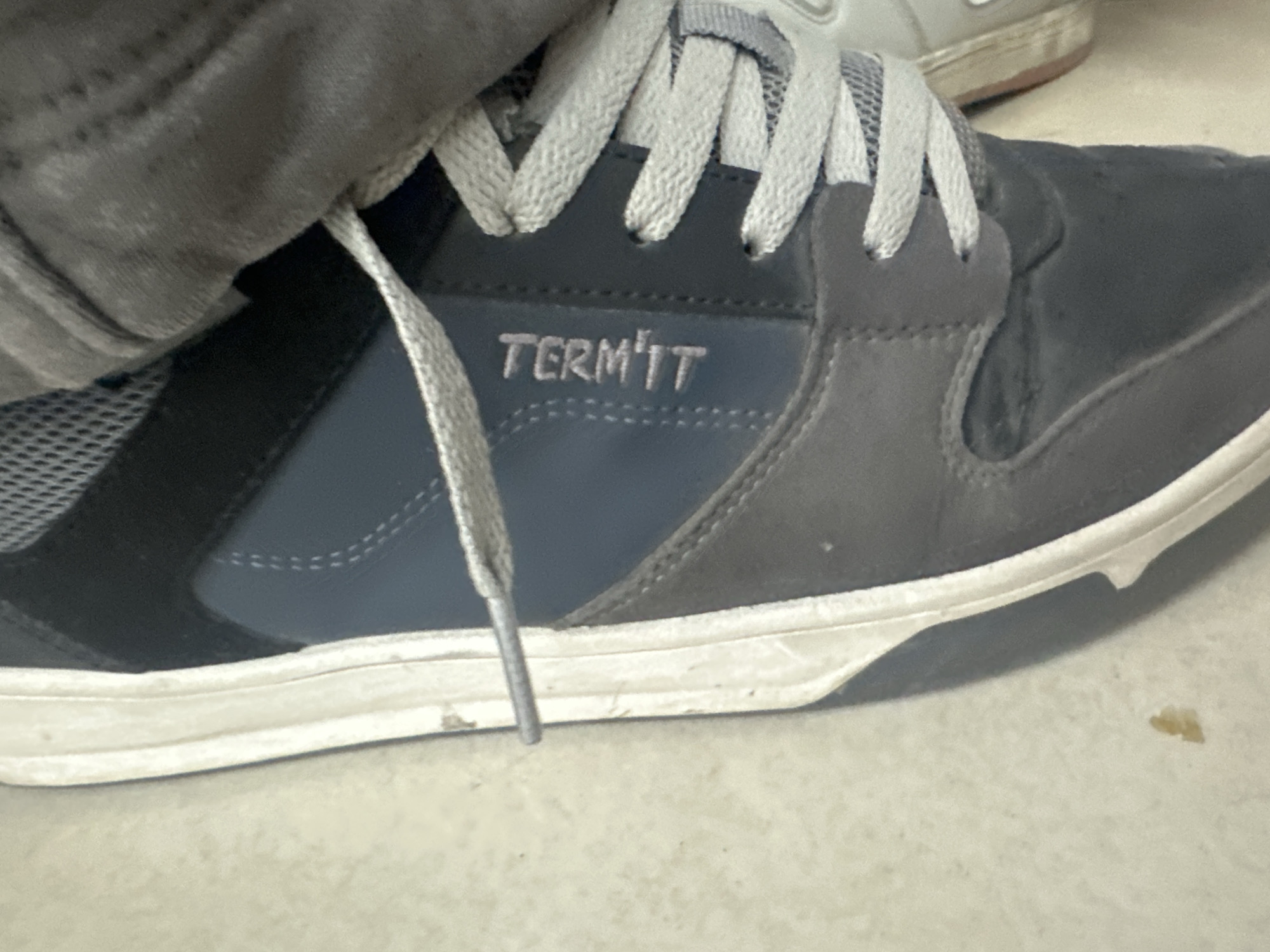 A close-up, eye-level shot captures the side of a casual shoe resting on a light-colored floor indoors. The shoe, a sneaker or skate-style shoe, features panels of dark grey, dark blue, and black mesh. Light grey laces are tied through eyelets on the tongue of the shoe.
On the dark blue side panel, the brand name "TERM'IT" is visible in light grey, stylized text. The apostrophe in "TERM'IT" appears as a vertical line. Part of a dark grey pant leg or sock is visible over the top of the shoe, suggesting it is being worn. In the blurred background, another light-colored shoe with a white sole and a tan bottom is partially visible. The floor beneath the shoe is a light, neutral color, possibly concrete or tile, with a small yellowish piece of debris or stain near the toe of the shoe. The overall impression is a candid shot of footwear in an everyday indoor setting.