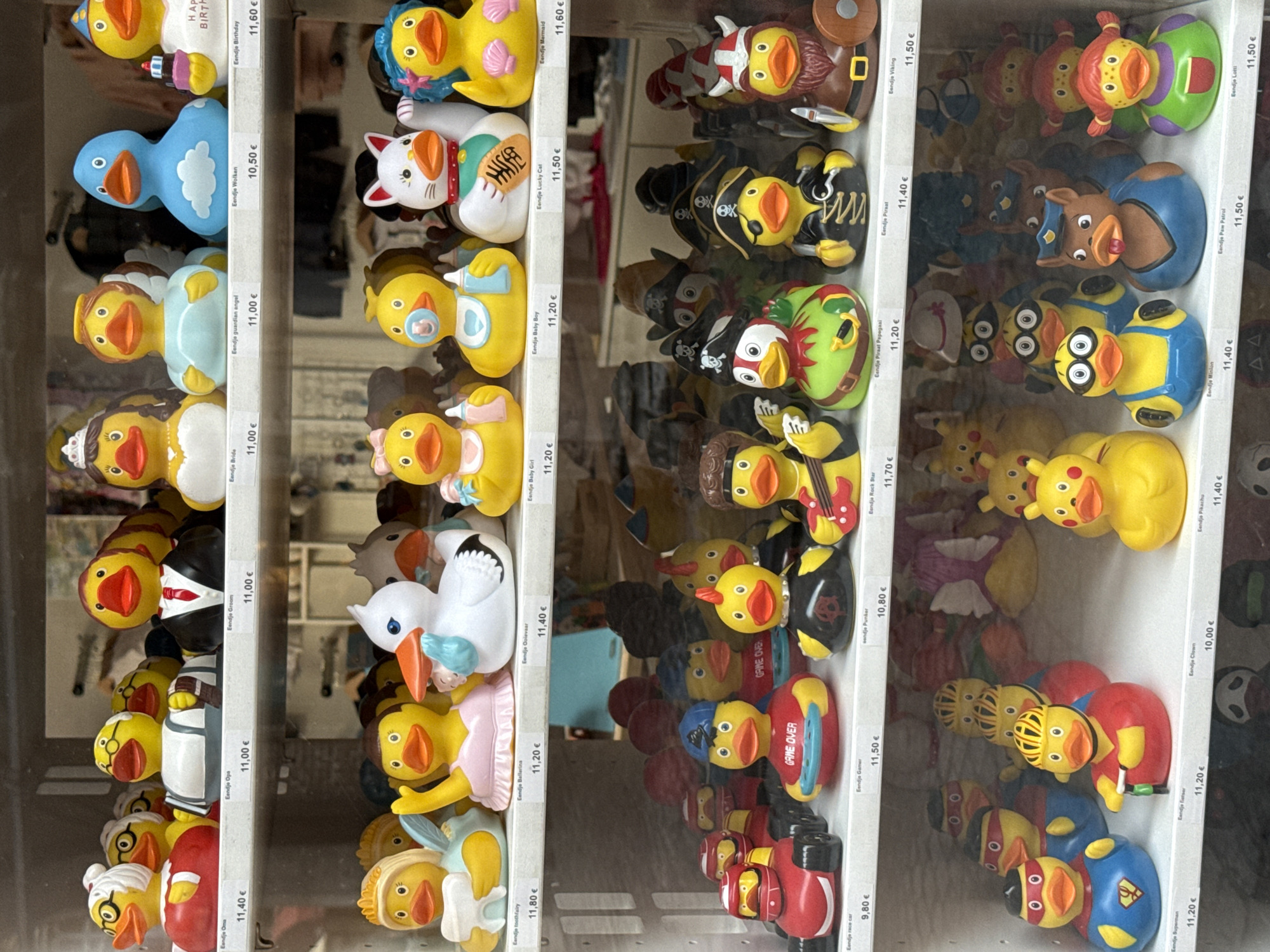This is a photograph taken inside a shop in Bruges, Belgium, exhibiting a collection of novelty rubber ducks displayed on shelves. The ducks are arranged in rows and are all facing forward. The first shelf displays a variety of character-themed ducks, including a bride, a guardian angel, a blue duck with clouds, and a birthday duck with a cake. The second shelf features more themed ducks such as a tooth fairy, a ballerina, a stork, a baby duck, a lucky cat, and a mermaid. The third shelf showcases a collection of ducks dressed as a race car driver, a gamer, a punker, and a rock star. The fourth shelf presents a group of ducks in superhero costumes, a cyclist, a clown, and a Pikachu character. The bottom shelf displays more themed ducks including a Minion, a police dog, and a character named Lotti. The ducks vary in color, design, and size, with some adorned with hats, costumes, and accessories. The background is blurred, suggesting the focus is on the merchandise. The lighting is bright, illuminating the colorful display of rubber ducks.