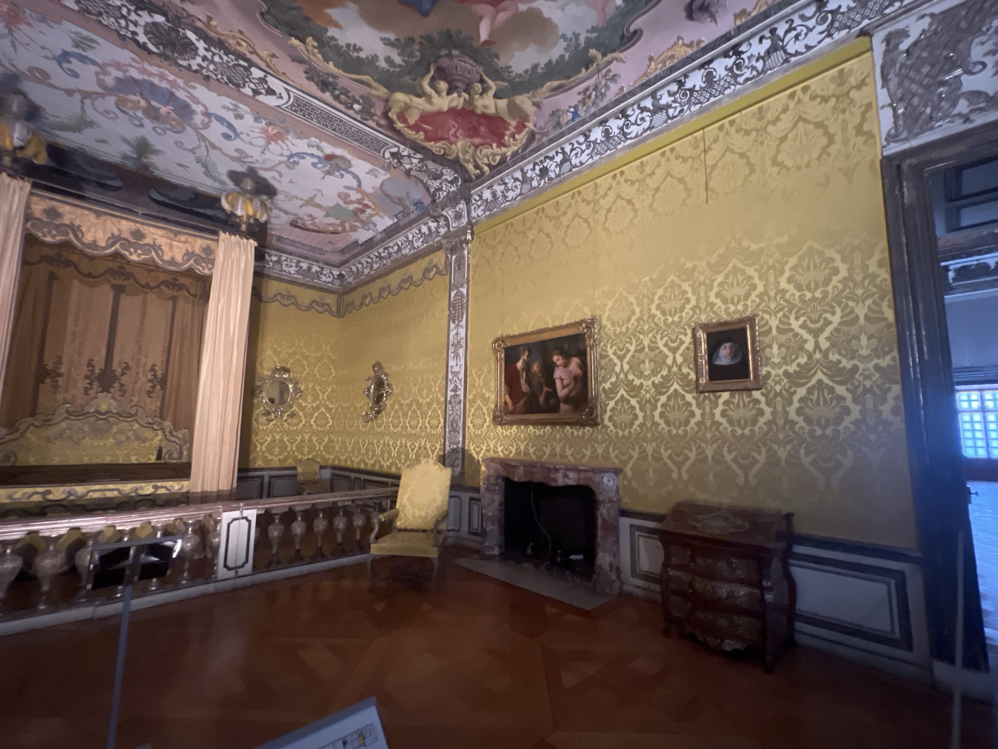 The image depicts a lavishly decorated room within the New Palace in Oberschleißheim, Germany. The room features an ornate frescoed ceiling with mythological scenes. The walls are covered in a textured golden damask wallpaper. On the left side of the room, a raised, curtained bed or alcove is visible, with a decorative railing in front of it. In the center of the room, there is a marble fireplace with a painting hanging above it, depicting a religious scene. To the right of the fireplace, a yellow armchair is placed. Further to the right, a dark wooden chest of drawers with decorative inlays stands against the wall. The floor is made of polished dark wood with a parquet pattern. The lighting appears to be artificial, casting a warm glow on the rich textures and colors of the room. The scene is static and devoid of any visible people.