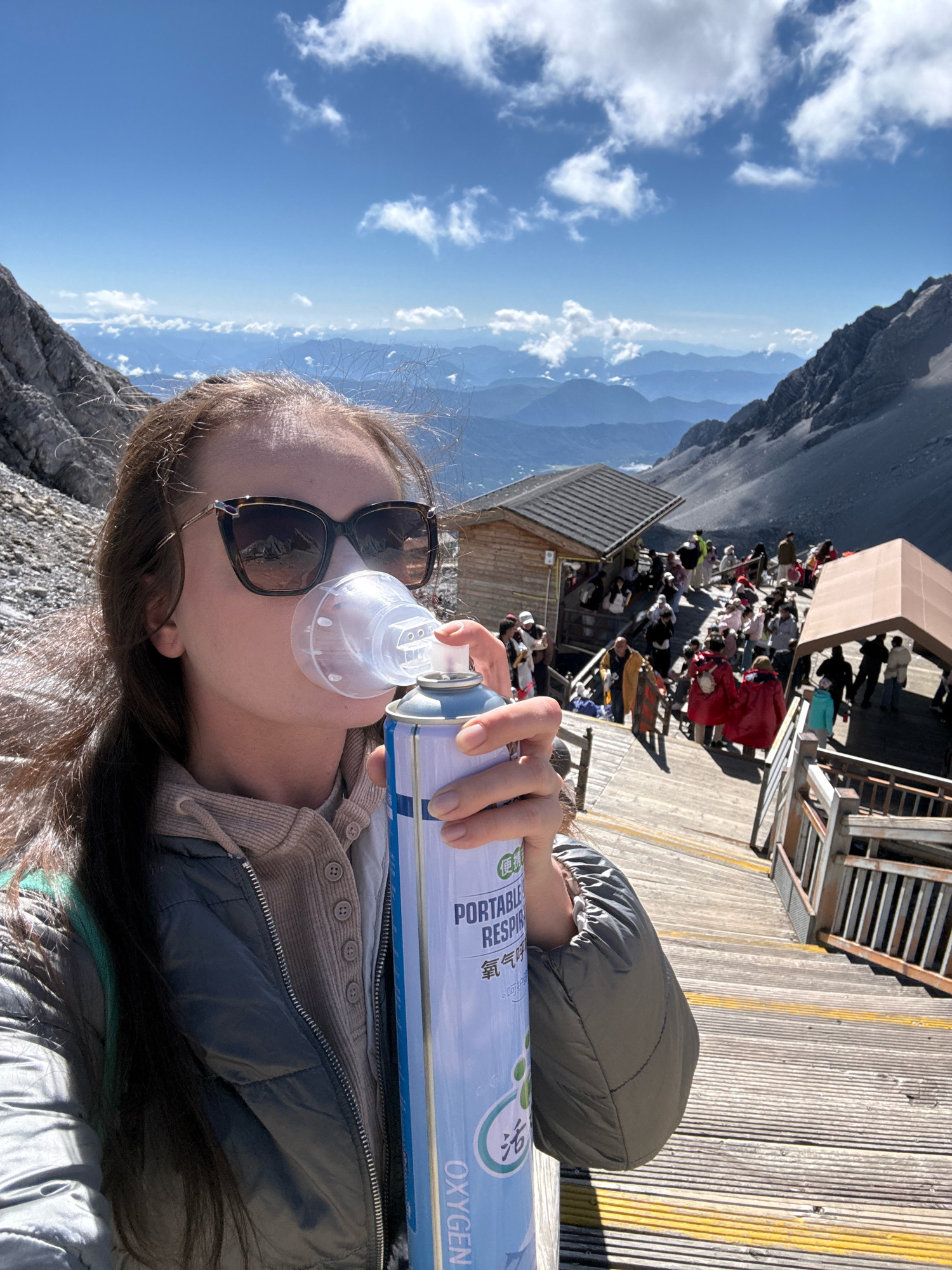 A woman is taking a selfie while using a portable oxygen canister at a high-altitude tourist attraction in Yulong Nakhi Autonomous County, China. The woman is wearing sunglasses and a gray jacket. She is holding a blue and white oxygen canister to her face, with a clear mask covering her nose and mouth. In the background, a wooden walkway leads up a steep incline, with numerous tourists walking and standing. Buildings with wooden roofs are visible on either side of the walkway. The environment is mountainous, with rugged terrain and distant peaks under a clear blue sky with scattered clouds.
