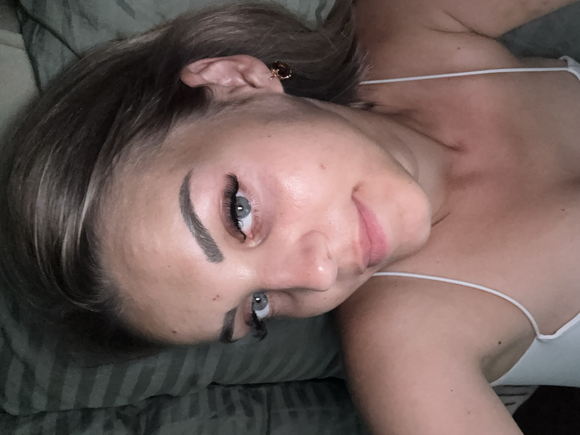This is a close-up, overhead, selfie-style shot of a young woman lying down, possibly in bed, given the presence of a striped pillow or blanket in the background. She has light brown hair with lighter streaks, styled loosely. Her eyes are a striking light blue, and she has dramatic false eyelashes. Her eyebrows are well-defined and arched. She is wearing a thin white spaghetti strap top. A small, gold-colored earring with a dark stone is visible in her right ear. Her skin appears slightly flushed, and there are a few small moles visible on her face and chest. The lighting is dim, suggesting it might be nighttime or an indoor setting with low light. There is no indication of the city of Yuzhno-Sakhalinsk, Russia, in the image itself.