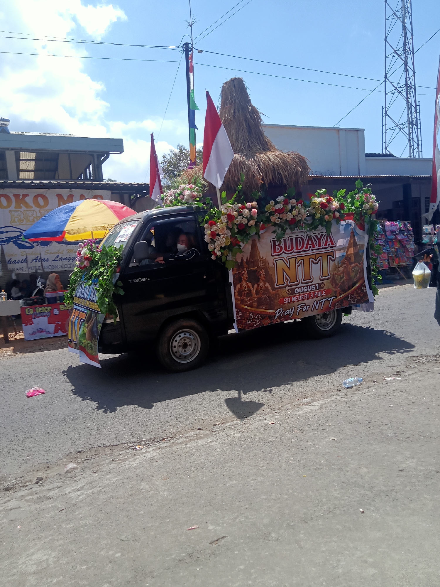 The image shows a black pickup truck decorated with flowers and banners driving on a road in the city of Pule, Indonesia. The truck is adorned with a large thatched headdress and two Indonesian flags. A banner on the side of the truck reads "BUDAYA NTT" and "Pray For NTT." A person wearing a face mask is visible driving the truck. In the background, there are buildings, a large telecommunications tower, and a blue sky with clouds. On the left side of the image, there is a colorful umbrella and some stalls selling goods. Several plastic bottles and wrappers are scattered on the road.