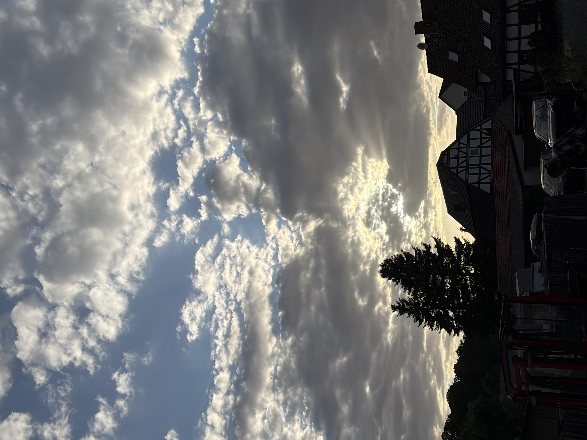 The image shows a cloudy sky over a village at dusk. The sky is filled with dramatic clouds, with patches of sunlight breaking through in the distance. Silhouetted against the sky are two tall pine trees. In the foreground, there are several buildings, including houses with traditional half-timbered architecture and red tiled roofs. There are also cars parked in front of the houses and a red tractor to the left of the frame. The overall impression is of a peaceful rural scene at the end of the day.