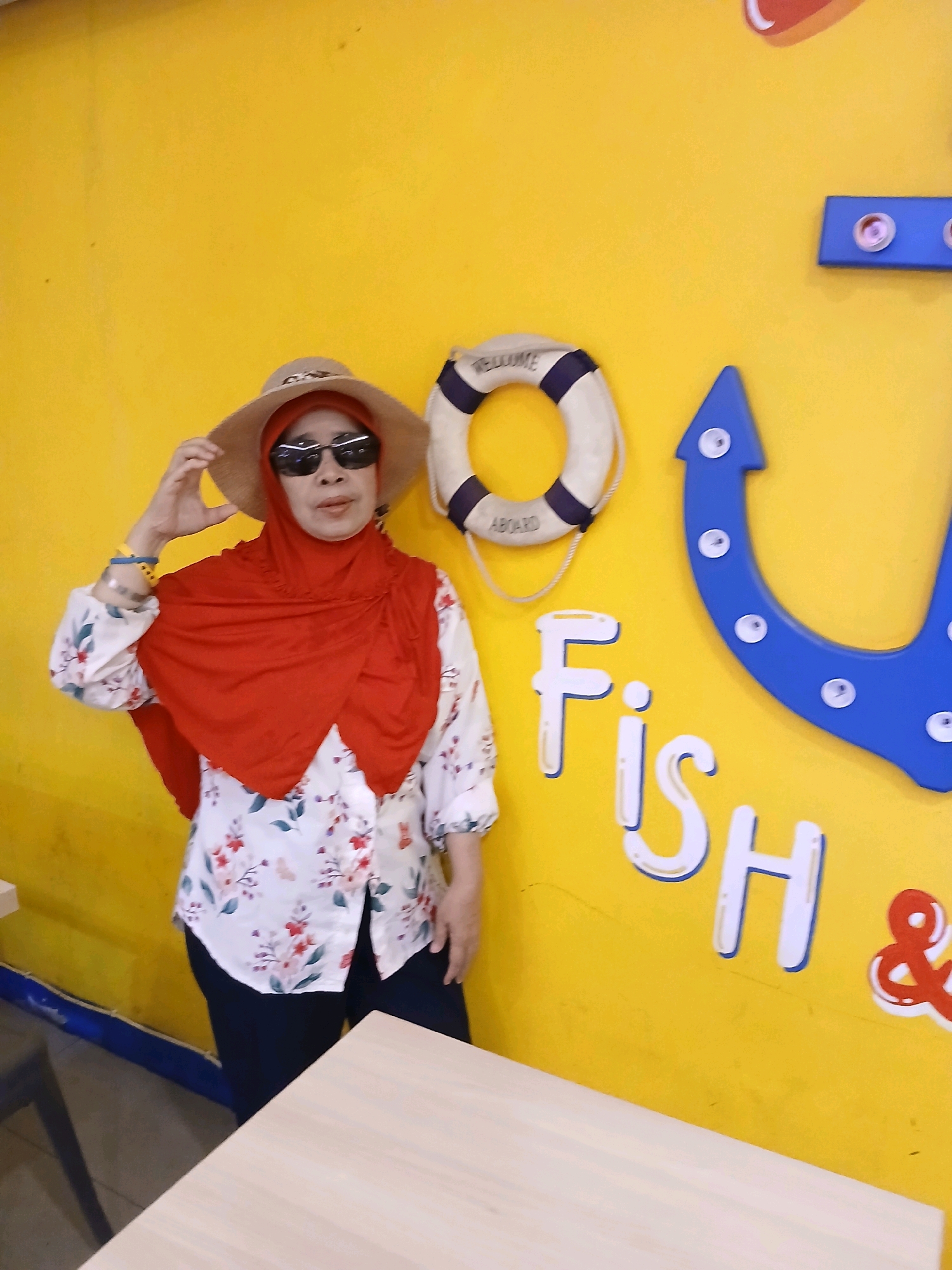 A woman wearing a red hijab, sunglasses, and a wide-brimmed hat stands in front of a bright yellow wall. She has a floral print shirt and dark pants. To her right, a life preserver hangs on the wall, and the word "Fish" is written in large white letters. Further to the right, a large blue fish-shaped decoration is visible. The scene appears to be an indoor decoration, possibly at a restaurant or recreational facility in South Jakarta, Indonesia.