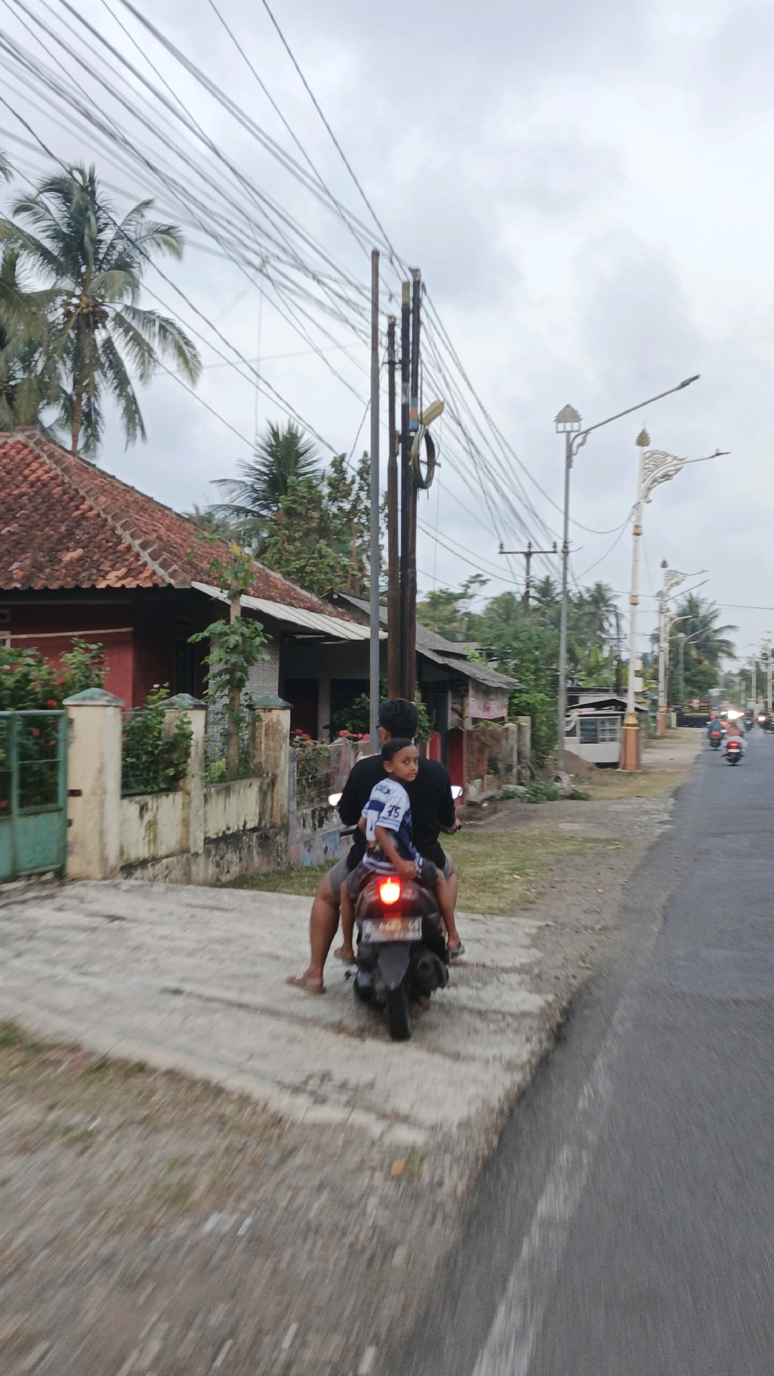 A father and son are riding a black motorcycle on a paved road in Desa Cijulang, Indonesia. The father is driving, wearing a black t-shirt and shorts. The son is sitting in front of him, wearing a blue and white striped shirt. They are both facing away from the camera. The motorcycle has a red taillight and a license plate that reads "B 4691 GS".
The environment appears to be a rural area with houses and palm trees lining the road. The sky is overcast, and there are numerous power lines strung across the scene. The road is asphalt, and there is a sidewalk or raised walkway made of concrete and gravel to the left of the road. In the background, several other motorcycles can be seen further down the road, suggesting traffic. The houses have red-tiled roofs and appear to be single-story structures. There are also decorative streetlights visible along the side of the road.
