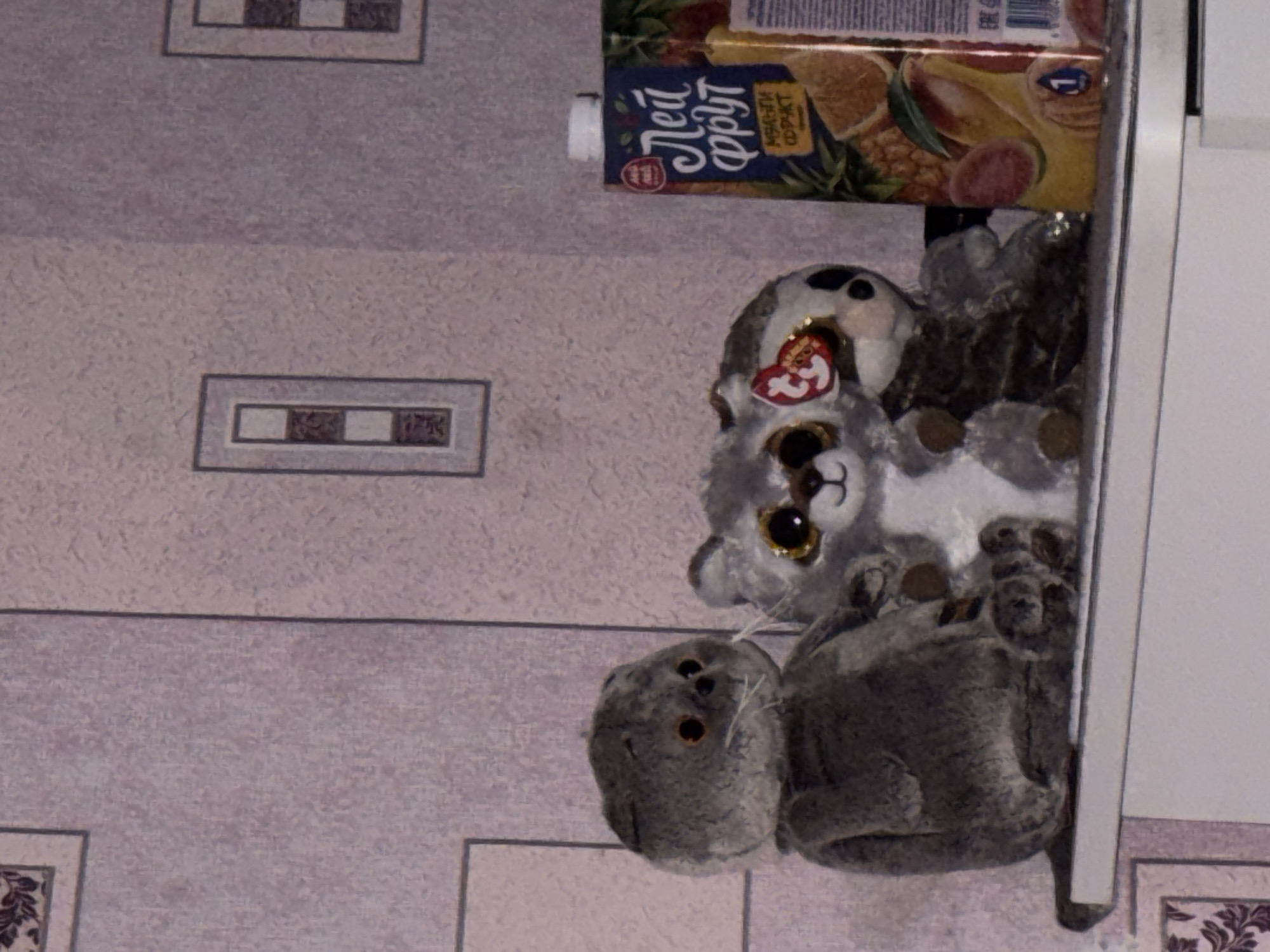 A close-up shot shows a carton of "Lei Fruit" juice resting on a cabinet surface next to several stuffed animals. The surface has a grey and white patterned design, possibly wallpaper or the cabinet's finish. The location is likely in Perm, Russia, given the Russian text on the juice carton. A grey cat stuffed animal is at the bottom, a grey and white animal is above and behind it, and a black and white animal is above and behind it. The carton of juice is at the top. The edge of the cabinet can be seen on the right side of the picture.