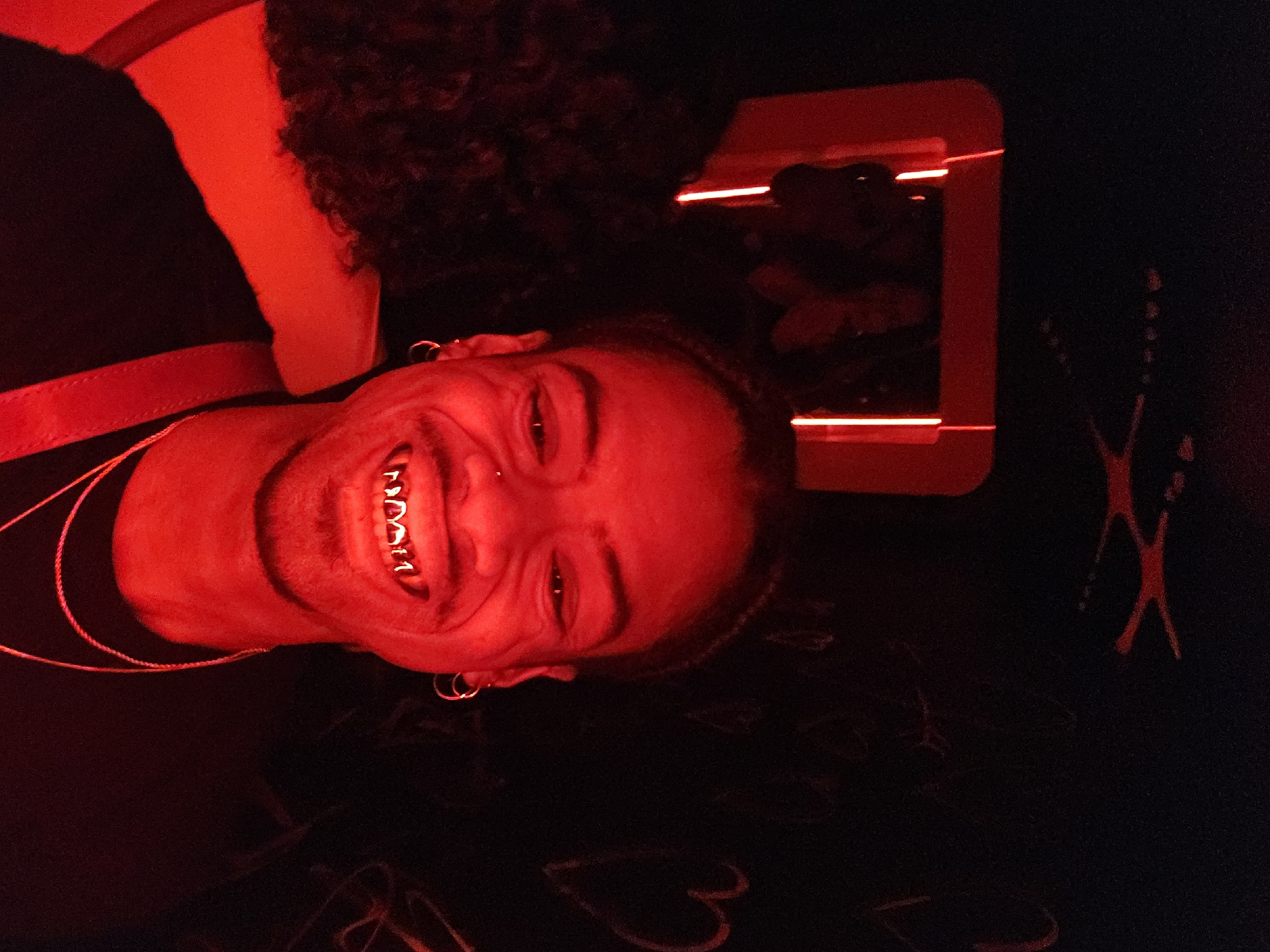 A person with dark curly hair and dark skin is illuminated by red light in a dark room. The person is smiling, revealing gold teeth. They are wearing a dark shirt and a thin gold chain. There is a red rectangular object with parallel light strips in the background. The background also includes white, graffiti-like markings of hearts, shapes, and an "X." The location cannot be determined from the image alone.