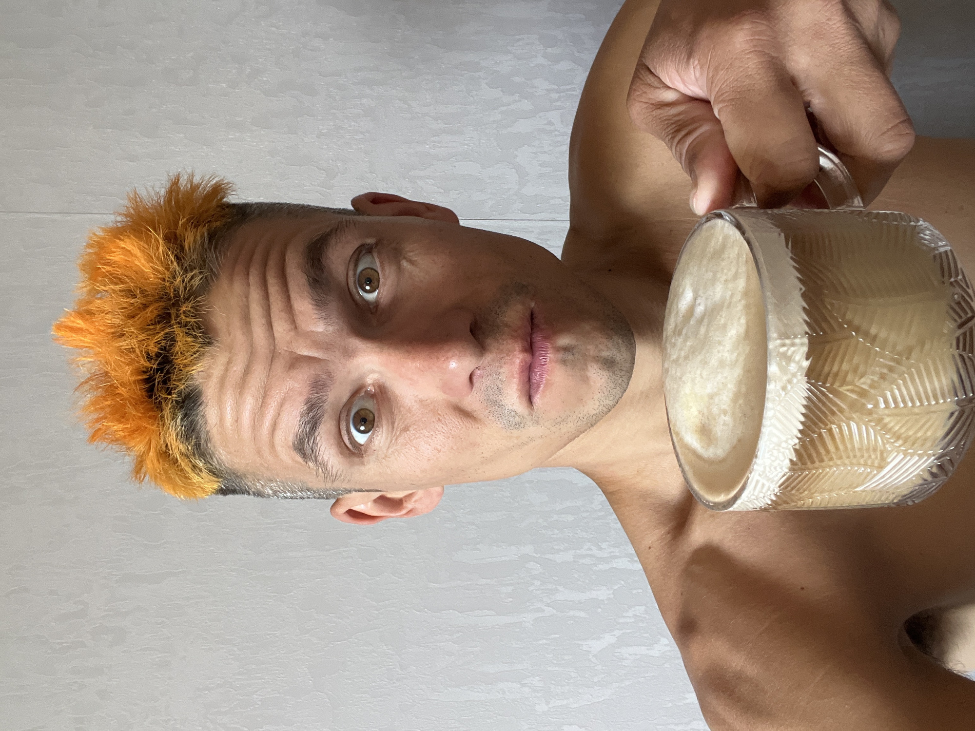A close-up, frontal, medium shot shows a shirtless man with spiky, bright orange dyed hair holding a clear glass mug filled with a frothy, light brown beverage. The man’s face is turned towards the camera, with his mouth slightly ajar and his eyes wide open, exhibiting a surprised expression. His skin appears tanned. The background is a plain, textured gray wall, suggesting an indoor environment. The man's right shoulder and arm are visible, as is his hand holding the mug. There is no discernible location in the city of Chaikovsky, Russia.