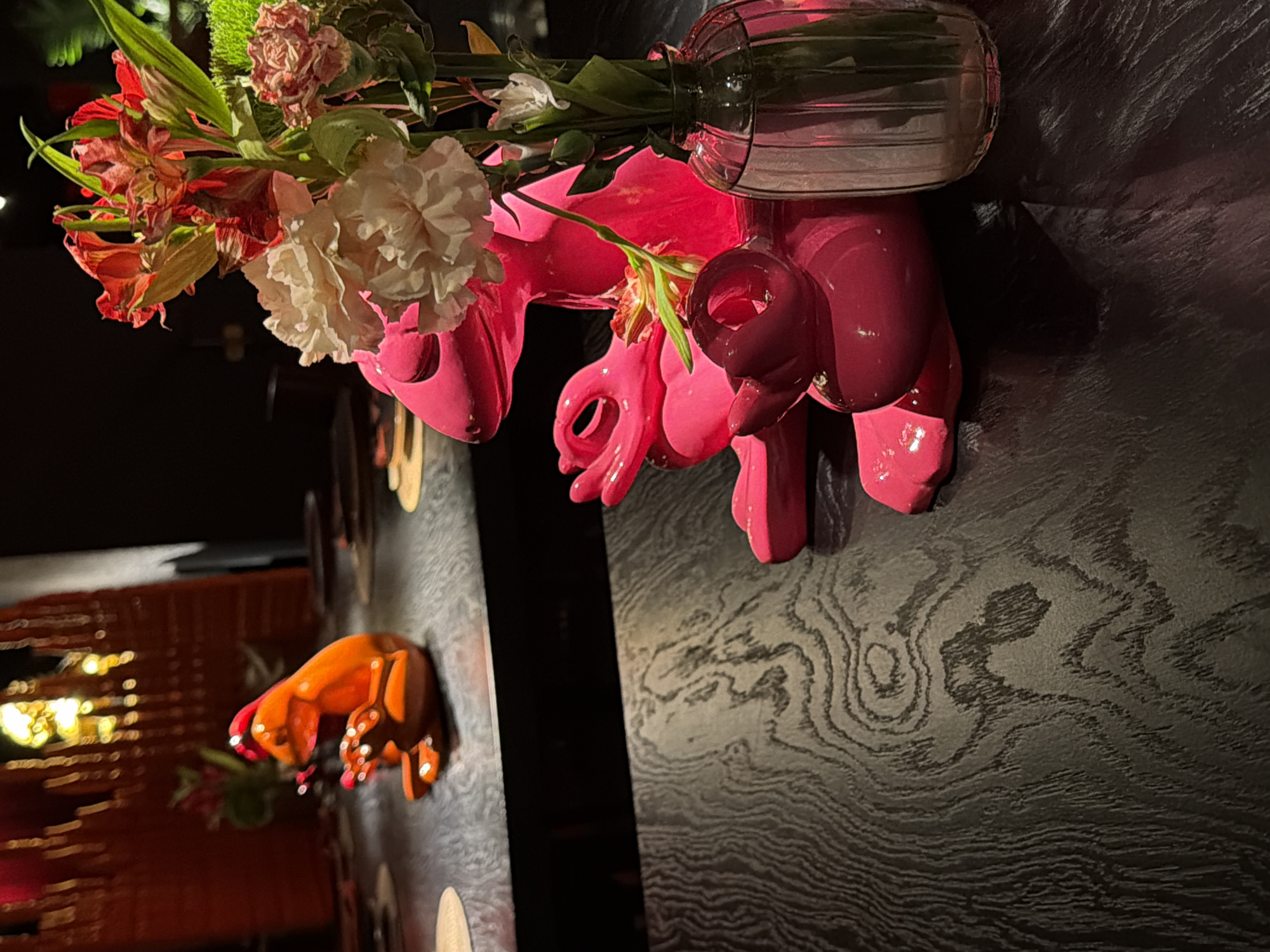 The image captures a decorative display on a table. A pink sculpture, resembling a whimsical animal, serves as a vase holding a bouquet of flowers in shades of red, green, and white. The vase appears to be made of clear glass. Further down the table, a similar sculpture in orange is also visible. The table itself has a dark, wood-grain finish. The lighting is dim, creating shadows and enhancing the colors of the objects. A blurred, warm-toned structure is seen in the background. The location is Moscow, Russia.