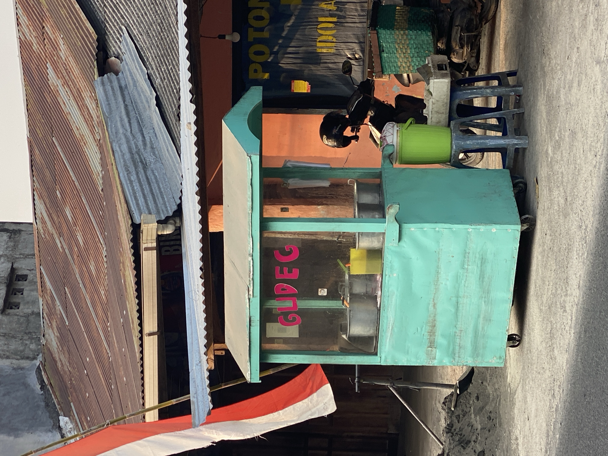 A street food stall with a bright teal wooden cart is set up in front of a building with a weathered, corrugated metal roof. The stall displays the word "GUDEG" in pink lettering on its glass window. Inside the cart, large metal pots and a yellow container are visible. To the right of the cart, a motorcycle is parked, and a black helmet rests on its handlebars. Two blue plastic stools sit on the ground, one supporting a green bucket and a white rectangular container. A woven green basket is on the back of the motorcycle. Behind the stall, an orange wall is visible, with a dark blue banner that has yellow lettering, partially obscured. A white and red Indonesian flag is draped on a pole to the left of the cart. The ground is paved with asphalt, and a pile of dark material, possibly sand or dirt, is to the left of the cart. The scene appears to be in the city of Kasihan, Indonesia, given the context of the food being sold.