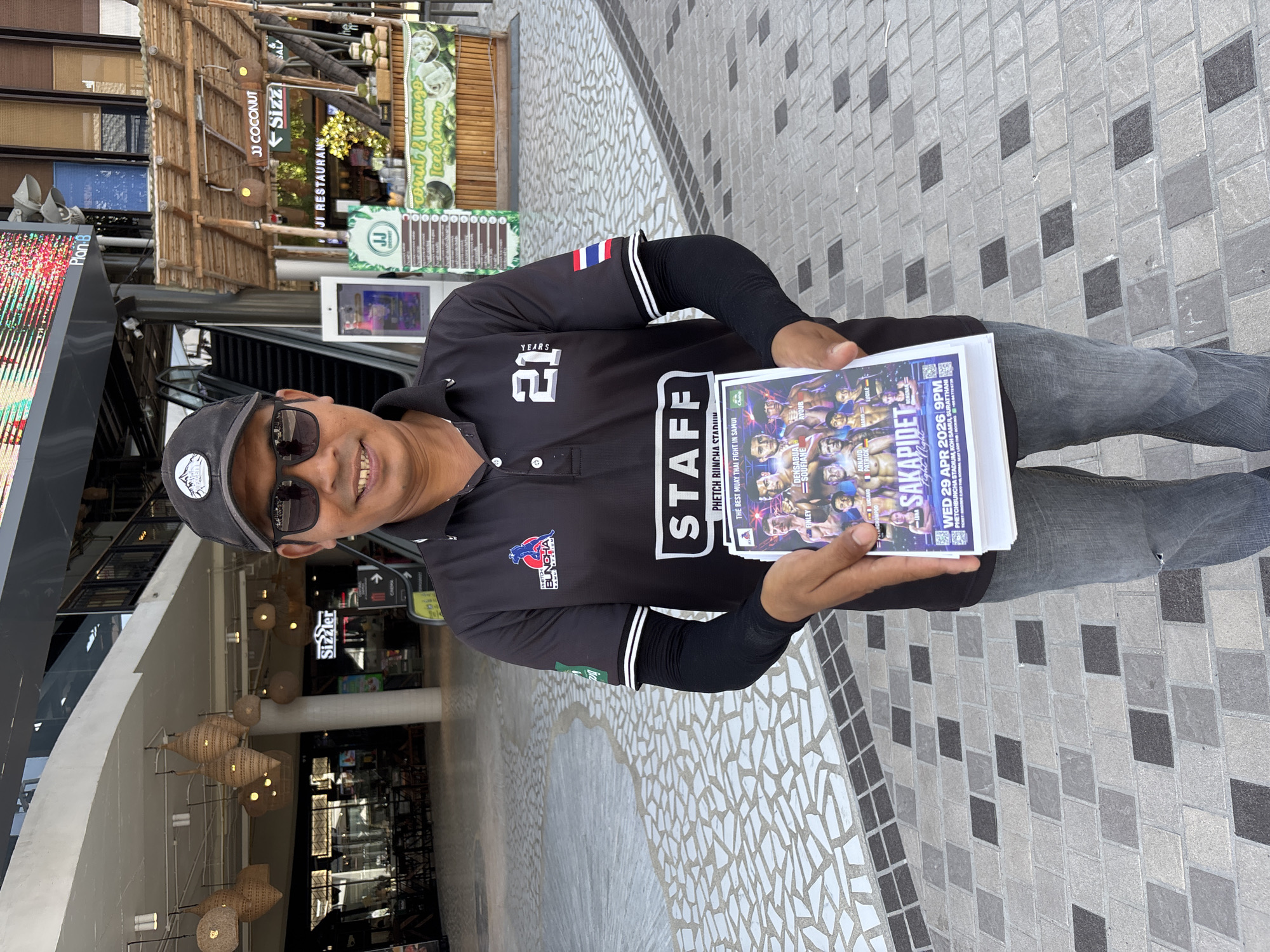 A man is depicted holding a stack of flyers and smiling at the camera, standing in what appears to be an outdoor commercial area or mall walkway. He is wearing a black short-sleeved polo shirt with "STAFF" and "21 YEARS" printed on it, along with a Thai flag on the sleeve and a "BUNCHA" logo. He also wears black arm sleeves, a black cap, sunglasses, and jeans.
The flyers he holds advertise a "SAKAPIDET Fight Night" at "PHETCHBUNCHA STADIUM, KOH SAMUI, SURATTHANI" on "WED 29 APR 2026 9PM", promoting it as "THE BEST MUAY THAI FIGHT IN SAMUI". Visible fighter names on the flyer include DENSABUA, SOUFIANE, AYOUB, ARNAUD PATRICK, LUCIANO, OMPOO, and LUCILE.
The background features various shopfronts and signs, including "JJ COCONUT", "Sizzler", "JI RESTAURANT", and a "Coconut & Mango Icecream" stand, some with bamboo-themed architectural elements. A large "Plan B" LED screen is visible on the left. The ground is paved with a distinctive pattern of light and dark gray square tiles. Decorative wicker-style hanging lamps are suspended from the ceiling in the mid-ground. The scene is brightly lit, suggesting daytime in Baan Chaweng, Thailand, where the event is likely being promoted.