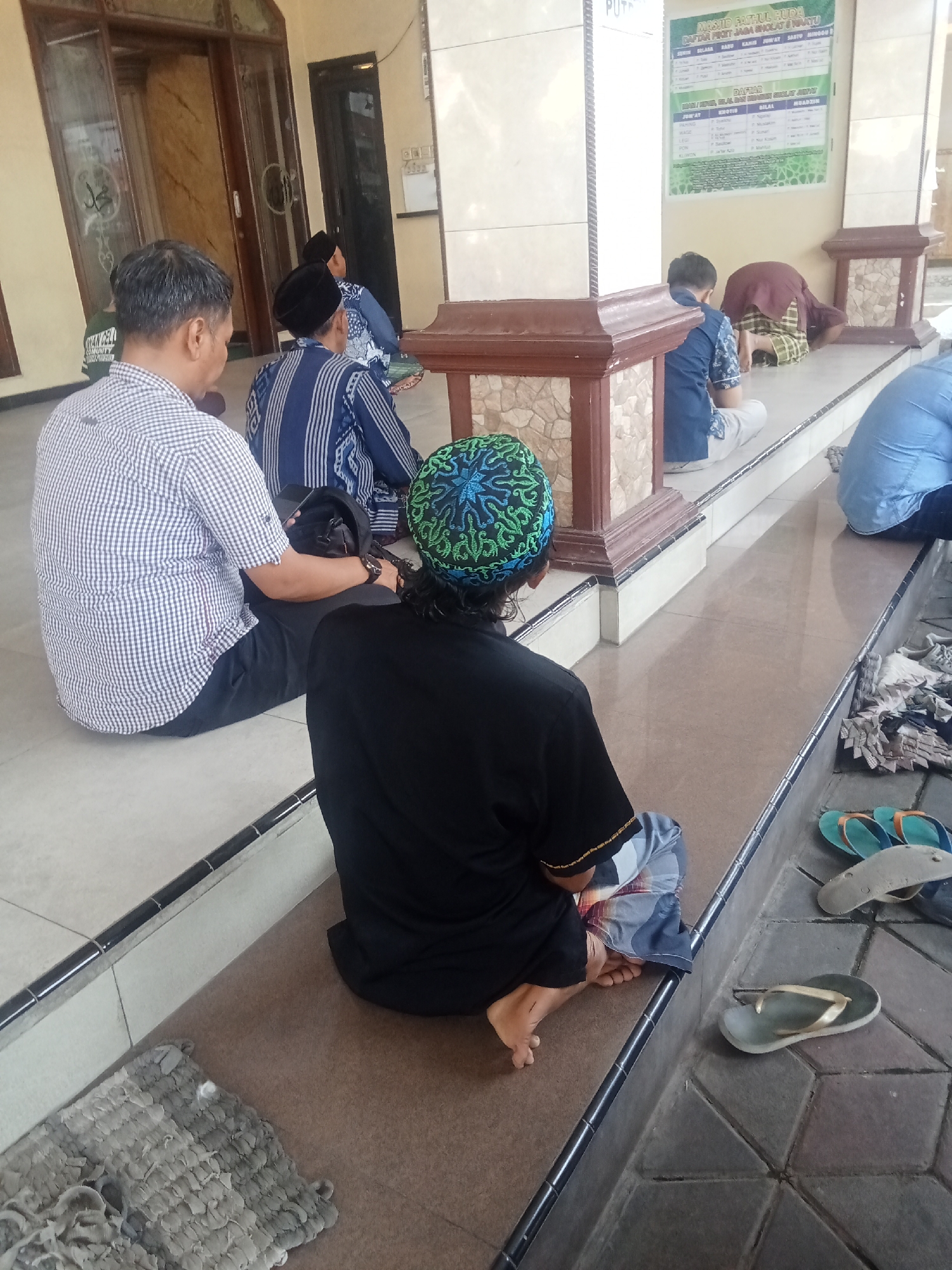 In the city of Menganto, Indonesia, a group of individuals are seen sitting on the tiled floor of what appears to be a mosque. The individuals are dressed in traditional Islamic attire, with several wearing kufi caps. In the foreground, a man wearing a checkered shirt is seated with his back to the camera, holding a bag. To his right, another man in a blue and white striped robe is also seated. Further in the background, a person is bowing down, presumably in prayer. Various pairs of flip-flops are scattered on the tiled walkway near the seating area. A poster with text and graphics is visible on a pillar in the background. The environment suggests a communal gathering for religious activities, possibly during a prayer service.