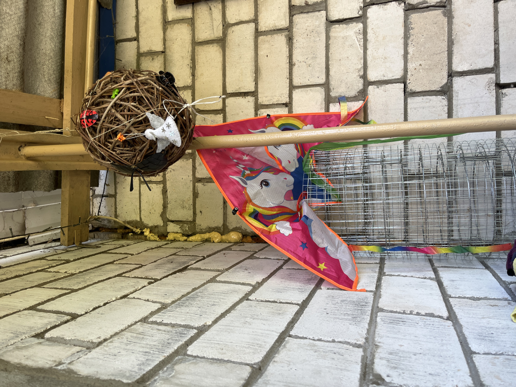 The image depicts an outdoor scene featuring a brick wall and hanging decorations. A large, roughly spherical ornament made of intertwined branches is suspended from a wooden beam. This ornament has several small, artificial insects attached to it, including ladybugs and possibly a spider. A white, felt angel ornament with glittery wings hangs from the same branch sphere. Below the branch sphere, a pink kite with a unicorn design is attached to a wooden pole. The kite has rainbow-colored streamers. The kite and pole are positioned in front of a wire mesh structure. The brick wall is constructed of light-colored cinder blocks with dark mortar. In the background, there appears to be a roof structure. The scene is illuminated by natural light, suggesting an outdoor environment. The location is identified as Дунілавіцкі сельскі Савет, Belarus.