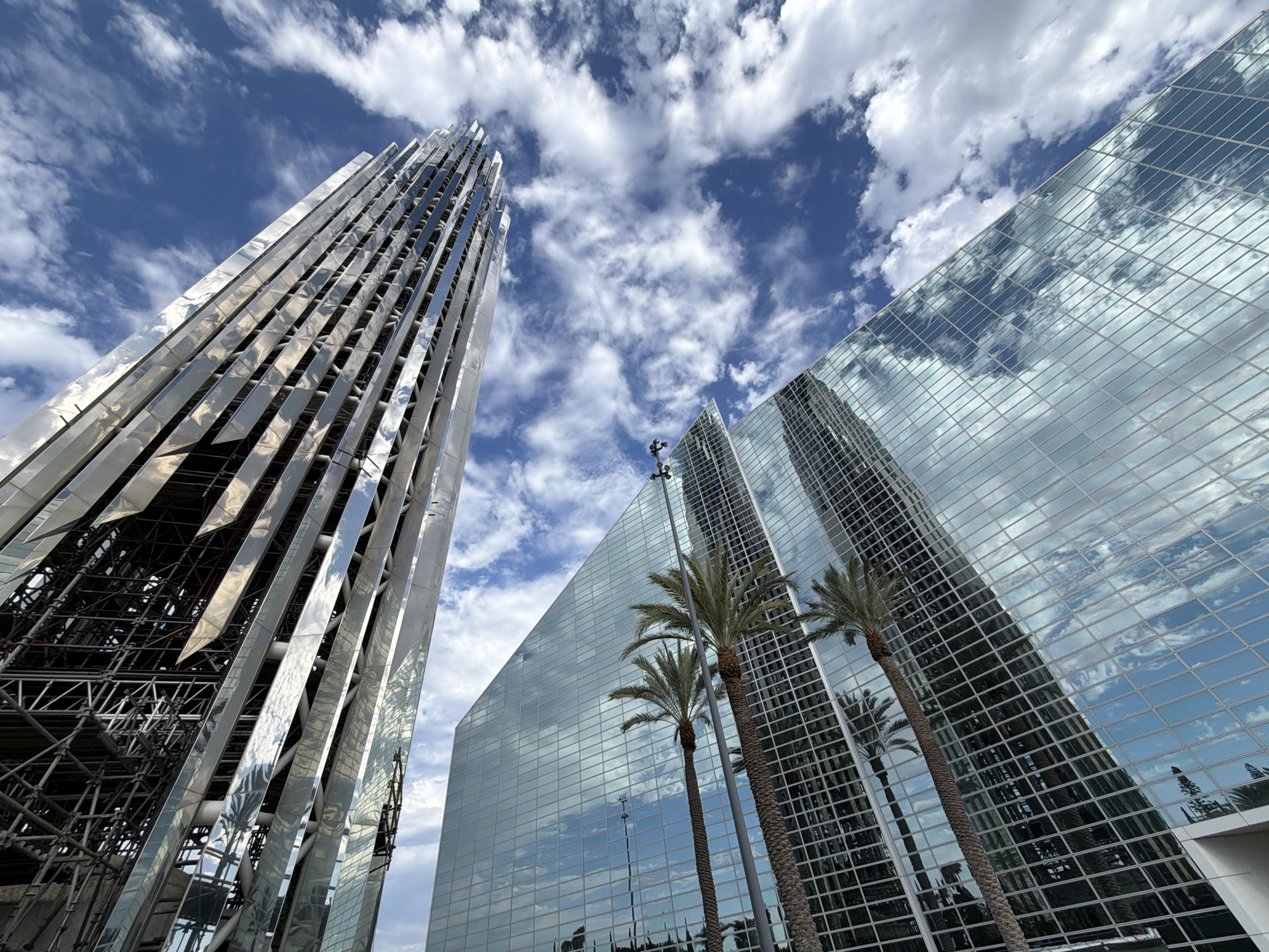 The image shows the exterior of a modern building complex in Garden Grove, California, United States of America. In the foreground, a tall, angular building with a glass facade reflects the sky and clouds. To the left, another structure, appearing to be under construction or renovation, features a metallic, ribbed exterior. Several palm trees are visible in front of the glass building, their reflections also captured in the facade. The sky is blue with scattered white clouds, indicating fair weather. The perspective is from a low angle, looking upwards at the buildings. There are no people visible in the scene.