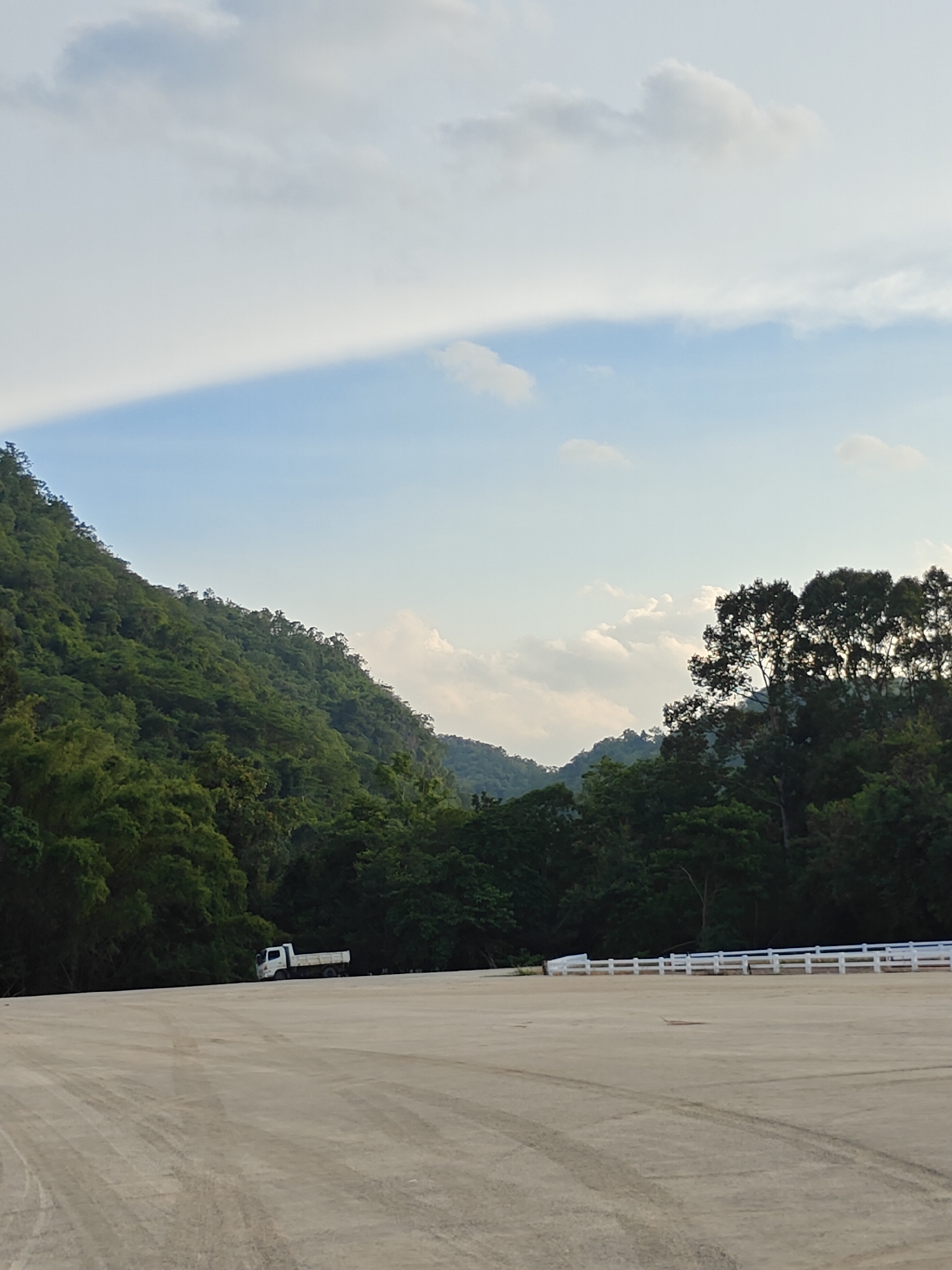 The image depicts a wide, flat, concrete area in Mu Si, Thailand, with tire tracks visible on the surface. In the background, a lush green, mountainous landscape dominates the scene, covered with dense vegetation. A white truck is parked in the middle ground, to the left of the frame. A white fence runs along the right side of the concrete area, separating it from the vegetation. The sky above is a mix of blue and white clouds, with a noticeable band of bright white cloud obscuring a portion of the sky. The lighting suggests it is daytime, likely late afternoon given the soft light.