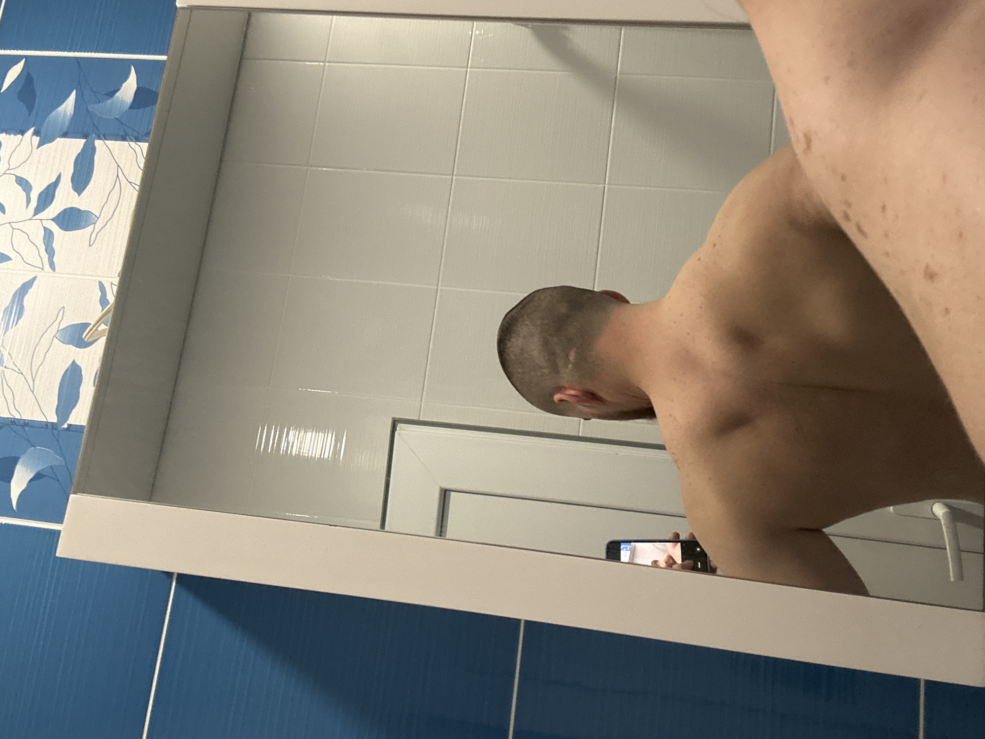 A mirror reflects a person with a shaved head and a bare back, holding a smartphone. The background reveals a tiled wall, with some tiles decorated with a blue and white floral pattern. The scene appears to be inside a bathroom or similar setting with a mirror and tiled walls.