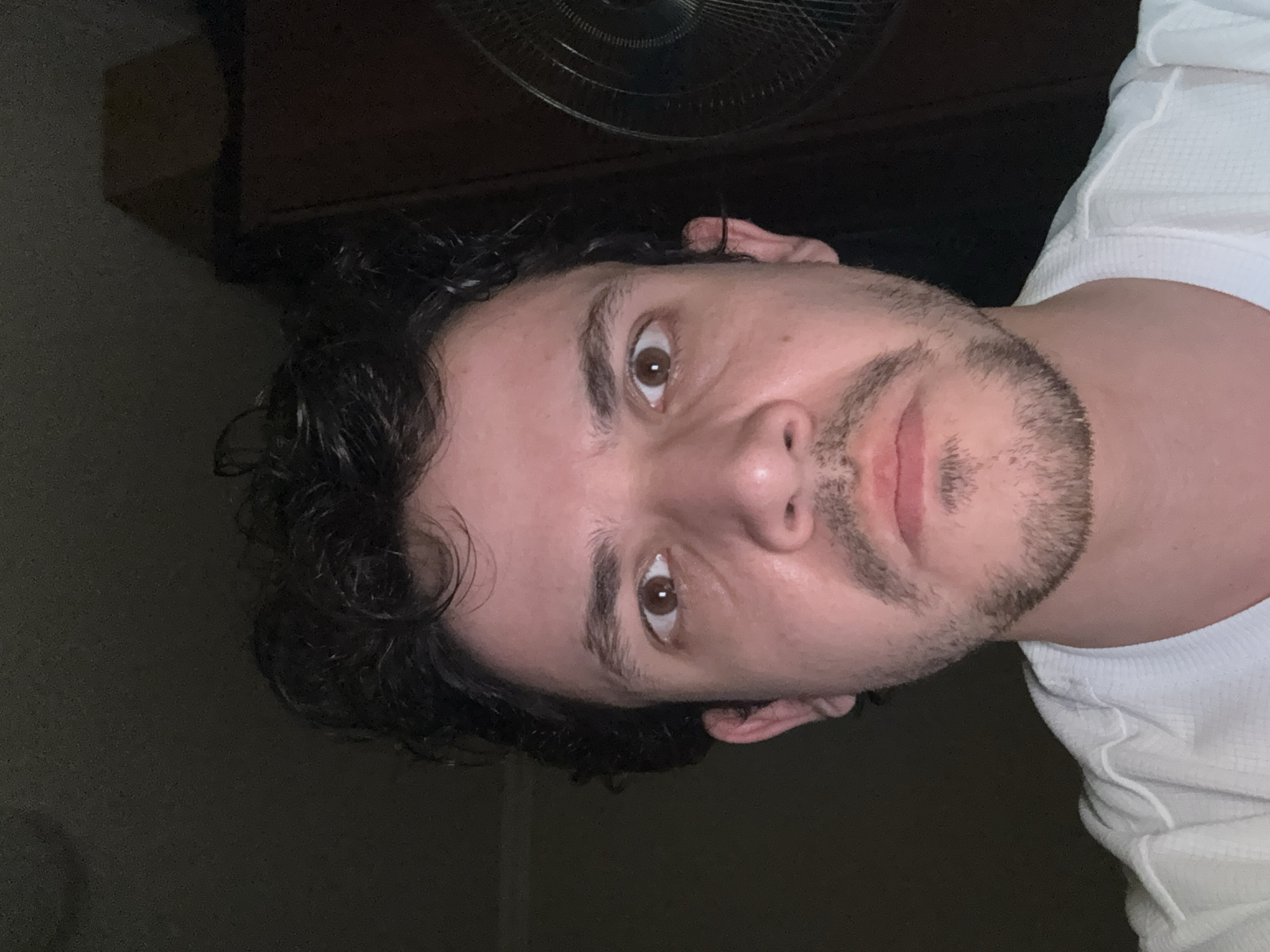 A man with dark, curly hair, brown eyes, and a short, dark beard is looking at the camera. He is wearing a white shirt with a waffle pattern. Above him, there is a fan with a dark background. The lighting is dim. It is impossible to tell from the picture if the image was taken in the city of Tashkent, Uzbekistan.