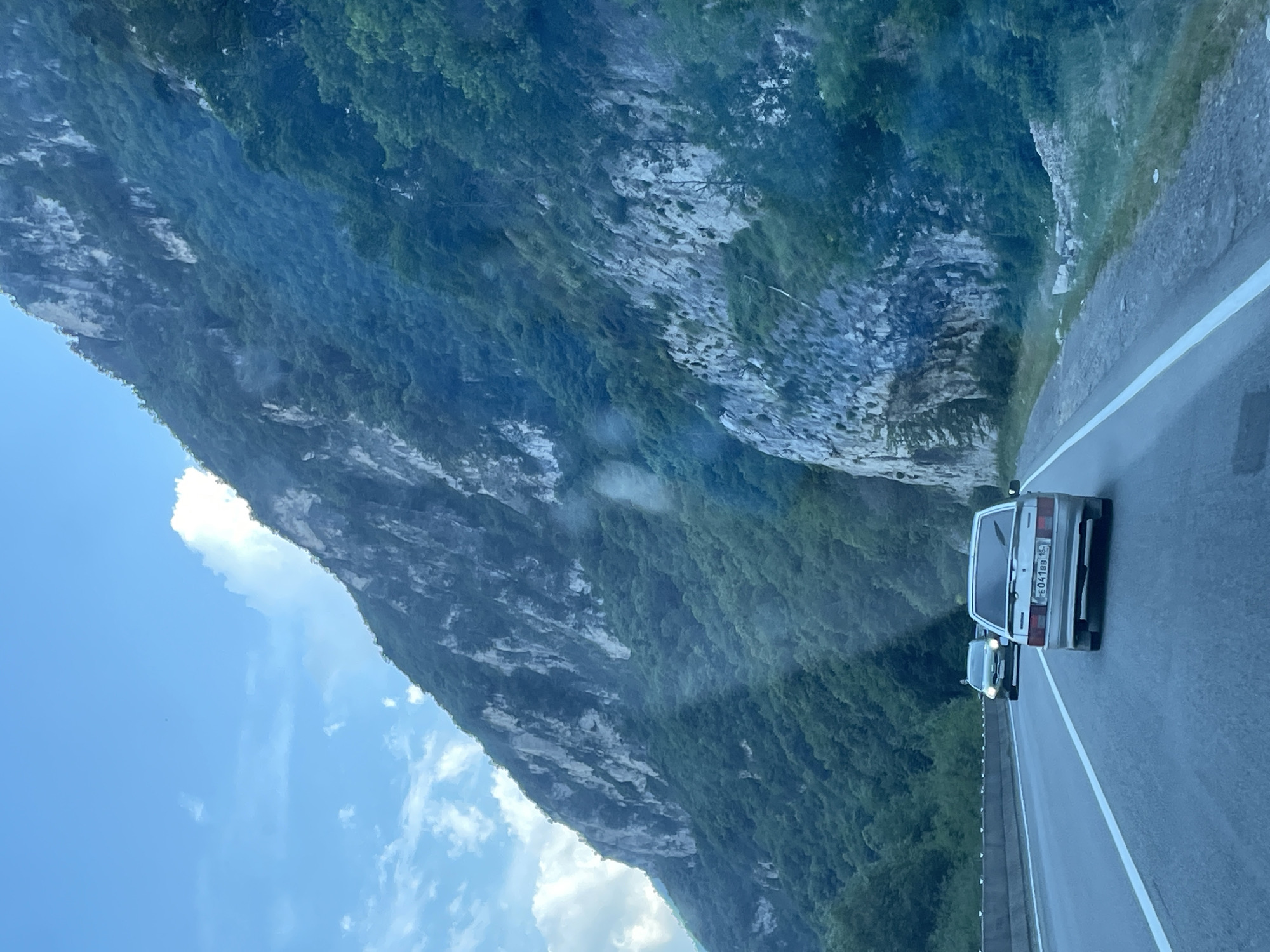 The image captures a scenic drive along a mountainous road, with lush green vegetation covering the slopes of a towering rock face. The sky above is a clear blue with a few scattered clouds. In the foreground, a silver car is driving on the asphalt road, with another car visible further ahead. The road appears to be a multi-lane highway, with white lines marking the lanes. The overall impression is one of natural beauty and a journey through a picturesque landscape. The location in the city of Balta, Russia is not explicitly mentioned in the image.