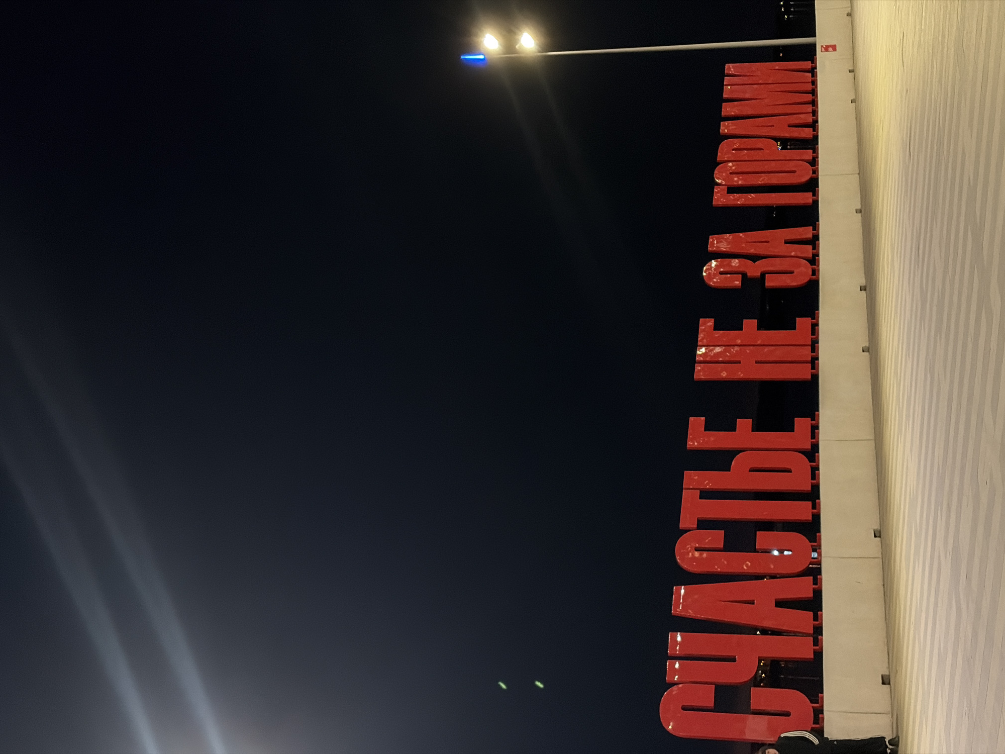 This is a nighttime photograph depicting the famous "Happiness is Just Around the Corner" art object in Perm, Russia. The scene is dominated by large, red, illuminated block letters spelling out the phrase "СЧАСТЬЕ НЕ ЗА ГОРАМИ" (Schastye ne za gorami), which translates to "Happiness is not far off" or "Happiness is just around the corner."
The text is prominently displayed horizontally, illuminated brightly against a dark, clear night sky that occupies the upper left portion of the image. To the right of the sign, a patterned, light-colored paved surface, likely a sidewalk or pedestrian area, extends into the background.
In the upper left corner, a metal pole extends, topped with a functional streetlight emitting a warm, bright glow, along with a smaller blue light. Rays of light from this streetlamp are visible against the dark sky. A person is partially visible at the bottom right corner, standing near the base of the sign, wearing dark clothing. The overall impression is that of a public space or landmark captured during the evening hours.
