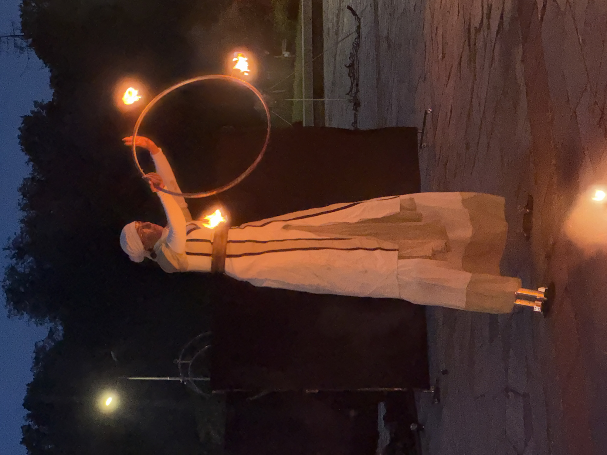 An outdoor night scene in Plovdiv, Bulgaria, features a fire performer on stilts. The performer, dressed in a light-colored tunic or robe with dark vertical stripes and a dark waist belt, also wears a matching light head covering. They stand on stilts, clearly visible at their feet, and are manipulating a metal hoop with three flaming wicks, two of which are actively burning brightly, casting warm orange light. The performer's gaze is directed towards the right.
In the background, dark silhouettes of trees are visible against a deep blue, twilight sky. A street lamp on a pole on the far left emits a bright yellow-white light. Behind the performer, a dark fabric backdrop or screen is erected, with metal support structures faintly visible. On the paved ground to the right of the performer, loose dark chains are scattered. The ground itself is composed of dark gray interlocking pavers or stones, with subtle reddish-orange illumination directly below the fire sources.