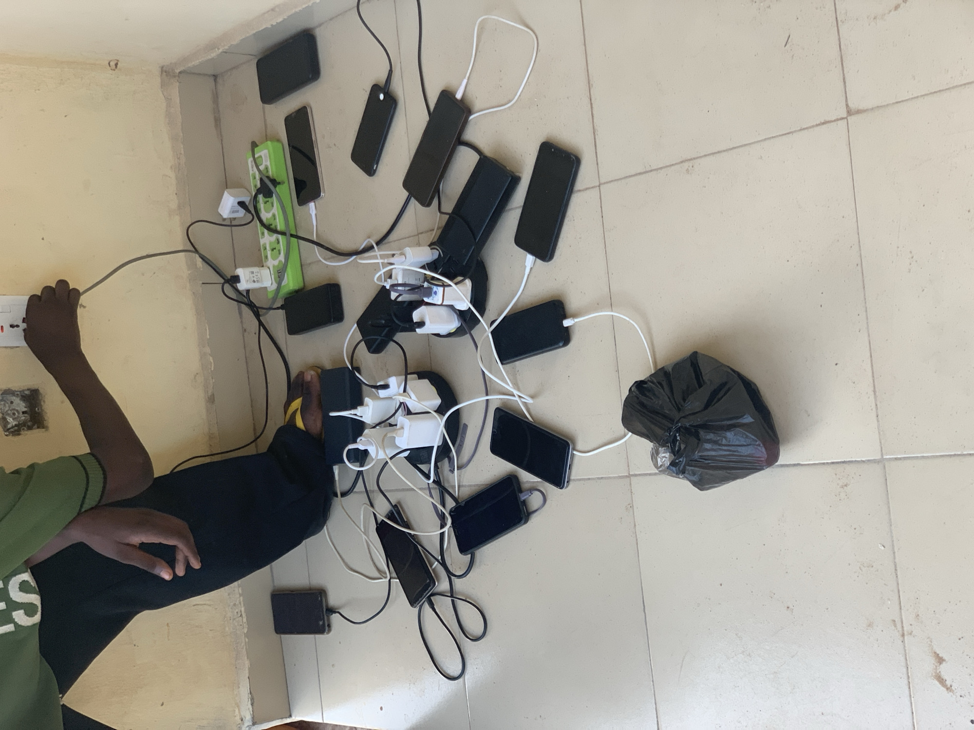 In an indoor scene in Lokoja, Nigeria, a person is seen managing an extensive setup for charging multiple mobile phones. The lower body and arm of the person are visible on the left side of the frame; they are wearing a green shirt with the partial text "ES" visible, black trousers, and yellow sandals. Their hand is near a white wall socket, which appears to be the primary power source.
From this wall socket, a grey cable connects to a green multi-socket power strip positioned against the wall. A complex array of black and white charging cables and adapters then branches out, connecting to numerous smartphones scattered across the light-colored, tiled floor. At least two large multi-port charging hubs, one green and one black, are visible, facilitating the connection of multiple white USB wall adapters.
Approximately 15 to 20 black or dark-colored smartphones are spread across the floor, each connected to a charger. The floor is light-colored and tiled, while the walls are a plain, light beige or yellow, showing some minor wear. A black plastic trash bag, tied at the top, rests on the floor to the right of the cluster of phones. The arrangement suggests a communal charging station or a scenario where many devices are charged simultaneously in one central location.