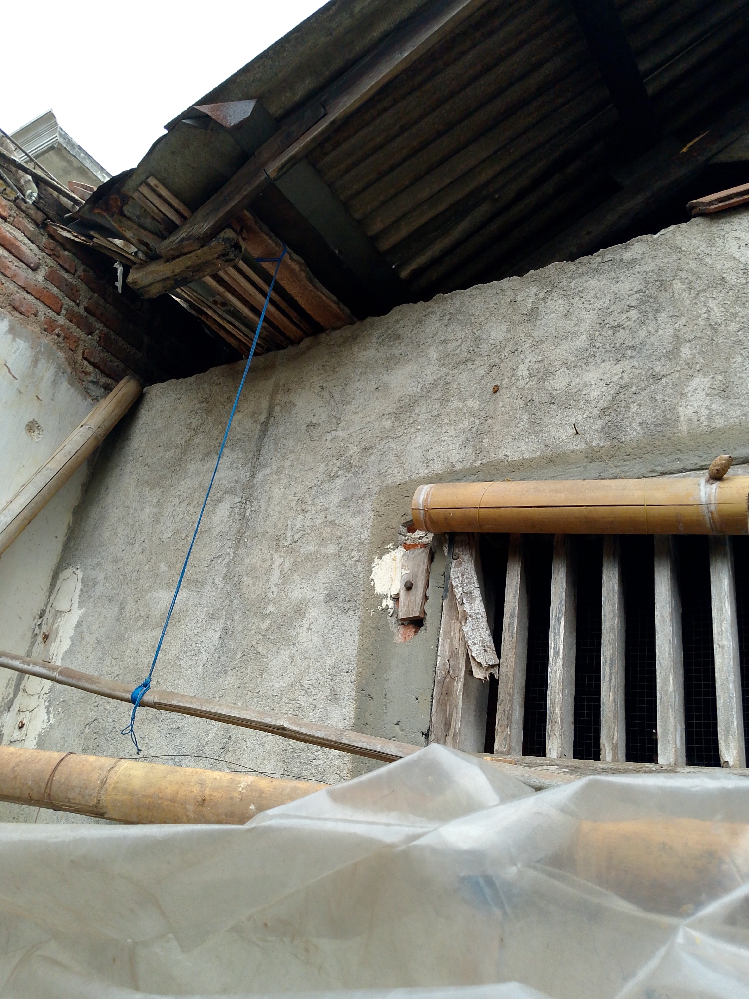 The image depicts a close-up view of a dilapidated structure, likely a building under repair or in disrepair. The foreground is dominated by a translucent plastic sheeting, partially obscuring what appears to be bamboo poles. Behind the plastic, more bamboo poles are visible, some horizontally positioned and others vertically, suggesting a framework or support system.

A textured concrete wall forms the central element of the image. A wooden structure with vertical slats, resembling a grille or window covering, is embedded in the wall. A horizontal bamboo pole is positioned across this grille. A blue rope is visible, running diagonally from the top left of the image, tied to a horizontal wooden beam, and extending downwards.

The upper part of the image reveals the underside of a damaged roof. Corrugated metal sheeting and weathered wooden planks are exposed, with some structural beams showing signs of decay. Exposed brickwork is visible on the left side, with crumbling plaster. The sky is a bright, overcast white. The scene suggests an outdoor environment in Babadan, Indonesia, characterized by rudimentary construction and weather-worn materials. No people are present in the image.