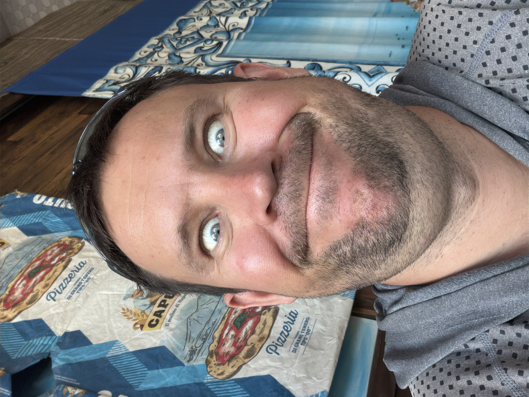In Baan Bo Phut, Thailand, a close-up, forward-facing selfie captures a man with wide, surprised eyes and a wide, goofy grin. He is wearing a gray hooded sweatshirt with a subtle diamond pattern. Behind him, a blue and white patterned fabric hangs, featuring images of pizzas and text. The fabric appears to be a decorative backdrop or possibly packaging. The lighting is bright, suggesting an outdoor or well-lit indoor setting. The man's facial expression dominates the frame, conveying a sense of playful amusement.