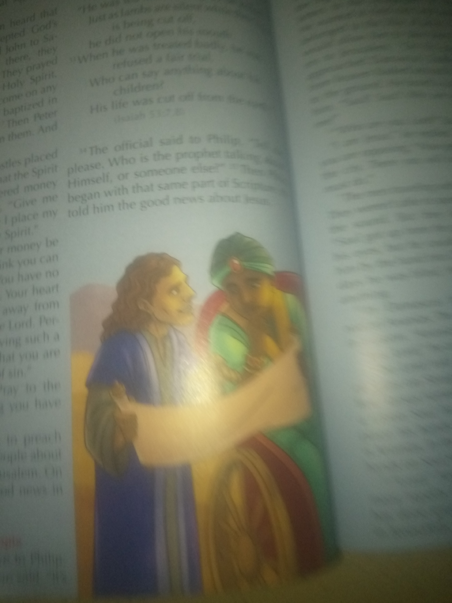 The image shows an open book with illustrations. The left page has text that appears to be from a religious text, with phrases like "n heard that," "epted God's," "baptized in," and "Holy Spirit." There is also a question that reads, "Who can say anything about your children?" and a statement, "His life was cut off from the earth." Further down, there is a quote, "The official said to Philip, 'Please, Who is the prophet talking about Himself, or someone else?' " and another sentence, "began with that same part of Scripture, told him the good news about Jesus."
The right page of the book contains more text, but it is largely illegible due to the focus and quality of the image.
In the foreground, there are two illustrated figures. The figure on the left appears to be a man with wavy brown hair, wearing a blue garment. He is looking to his right and holding a scroll or a map. The figure on the right is wearing a green turban and a green and red garment. This figure is facing left and appears to be speaking or gesturing towards the scroll. They are standing against a blurred background that suggests an indoor setting or a stylized outdoor scene.
There is no information in the image that would suggest a location in the city of Jauro Yinu, Nigeria.