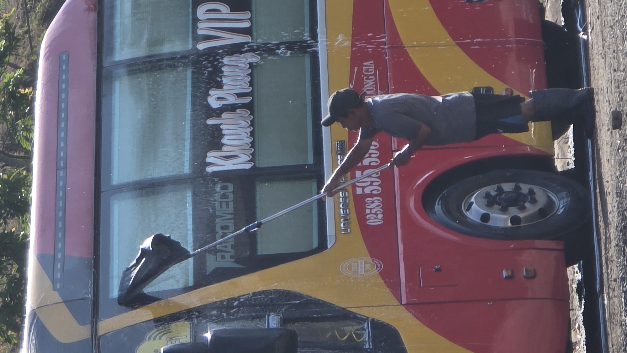 In Bắc Nha Trang, Vietnam, a man in a dark cap, light gray shirt, and dark shorts is washing a bus. The bus is red and yellow with white lettering, including "Khank Phong VIP" and a phone number, "02583 595 550". The man uses a long-handled squeegee to clean the bus windows. Soap suds are visible on the bus. The setting appears to be outdoors on a paved surface.