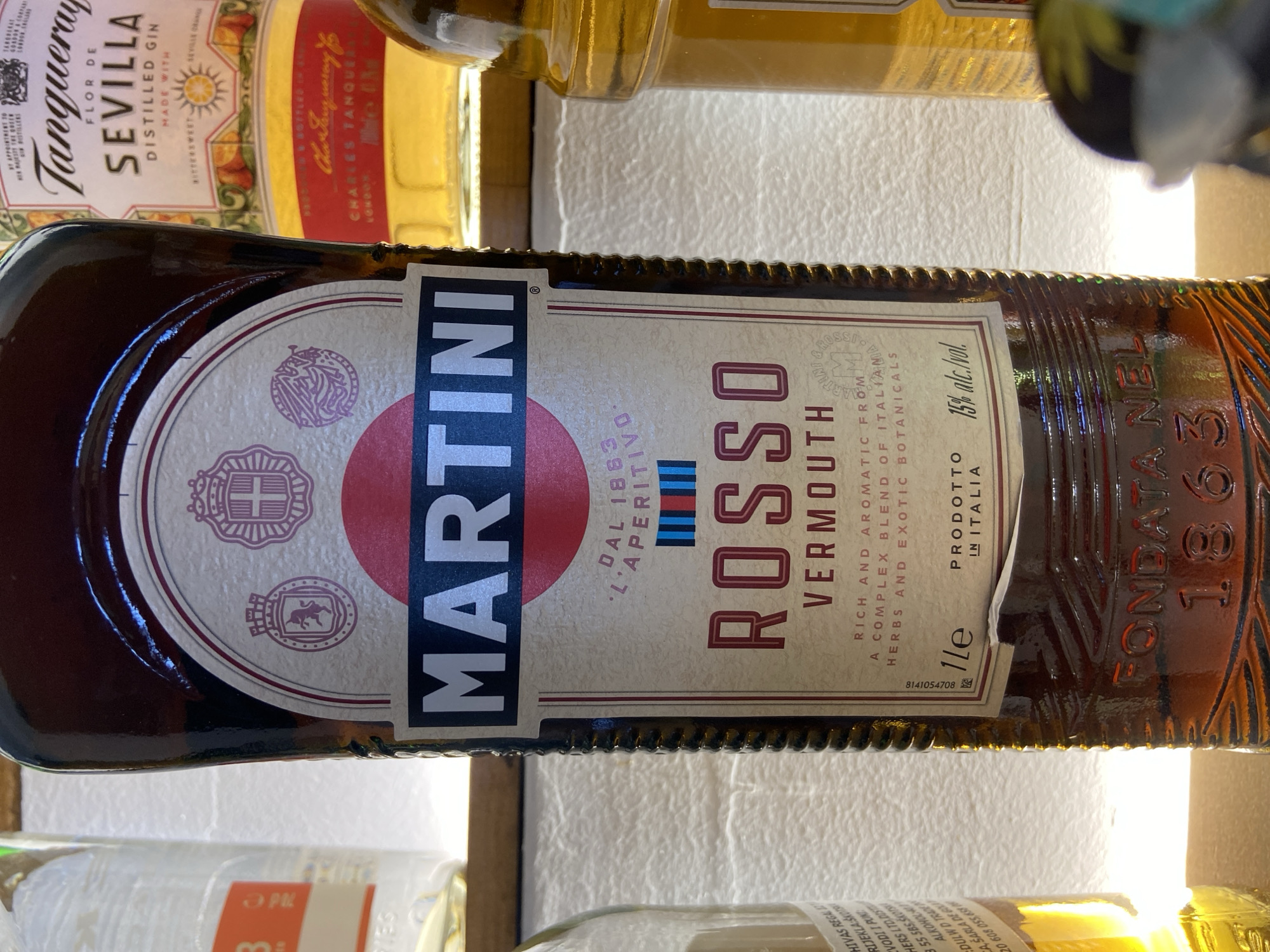 The image shows a bottle of Martini Rosso Vermouth, positioned in a location that appears to be a bar or shelf. Other bottles are partially visible. The Martini bottle is centered in the frame, with its label prominently displayed. The label includes the brand name "MARTINI," the product name "ROSSO VERMOUTH," and details about its flavor profile. A bottle of Tanqueray Sevilla gin is also visible in the background. The bottle is dark brown and has an intricately designed label. The environment appears to be well-lit.