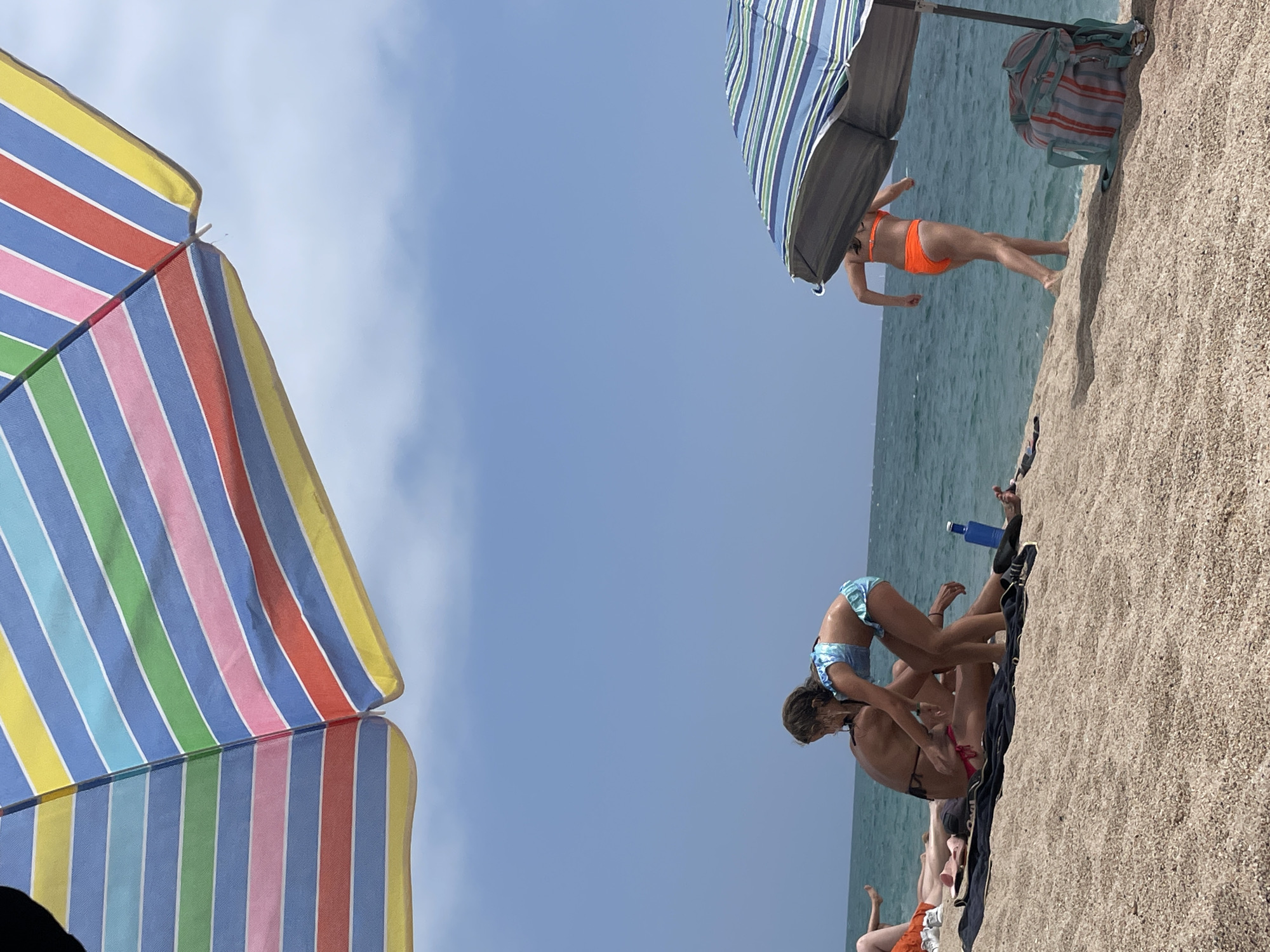 The media file depicts a beach scene in Canet de Mar, Spain, under a clear blue sky with some wispy clouds, suggesting a sunny day. Two large, colorful beach umbrellas, one with red, pink, blue, green, yellow, and white stripes and another with blue, white, and green stripes, are prominent in the foreground and midground.
Several people are engaged in activities on the sandy beach. In the midground, a person in an orange bikini is standing under the blue and green striped umbrella, appearing to be in motion, possibly dancing or stretching. Further to the right, on the sand, two individuals are interacting. One, wearing a blue patterned bikini, is in a playful pose, leaning over another person who is lying on a towel. A blue water bottle and a pair of black slip-on shoes are visible next to them. In the lower part of the frame, other beachgoers are partially visible, including someone in orange shorts.
The ocean is calm and a light blue-green, stretching towards the horizon. The sand appears to be a typical beach sand, with small shell fragments mixed in. The orientation of the image is rotated, with the sky appearing at the top and the sea at the bottom. The overall atmosphere is relaxed and indicative of a leisurely day at the beach. There is no visible text in the media file.