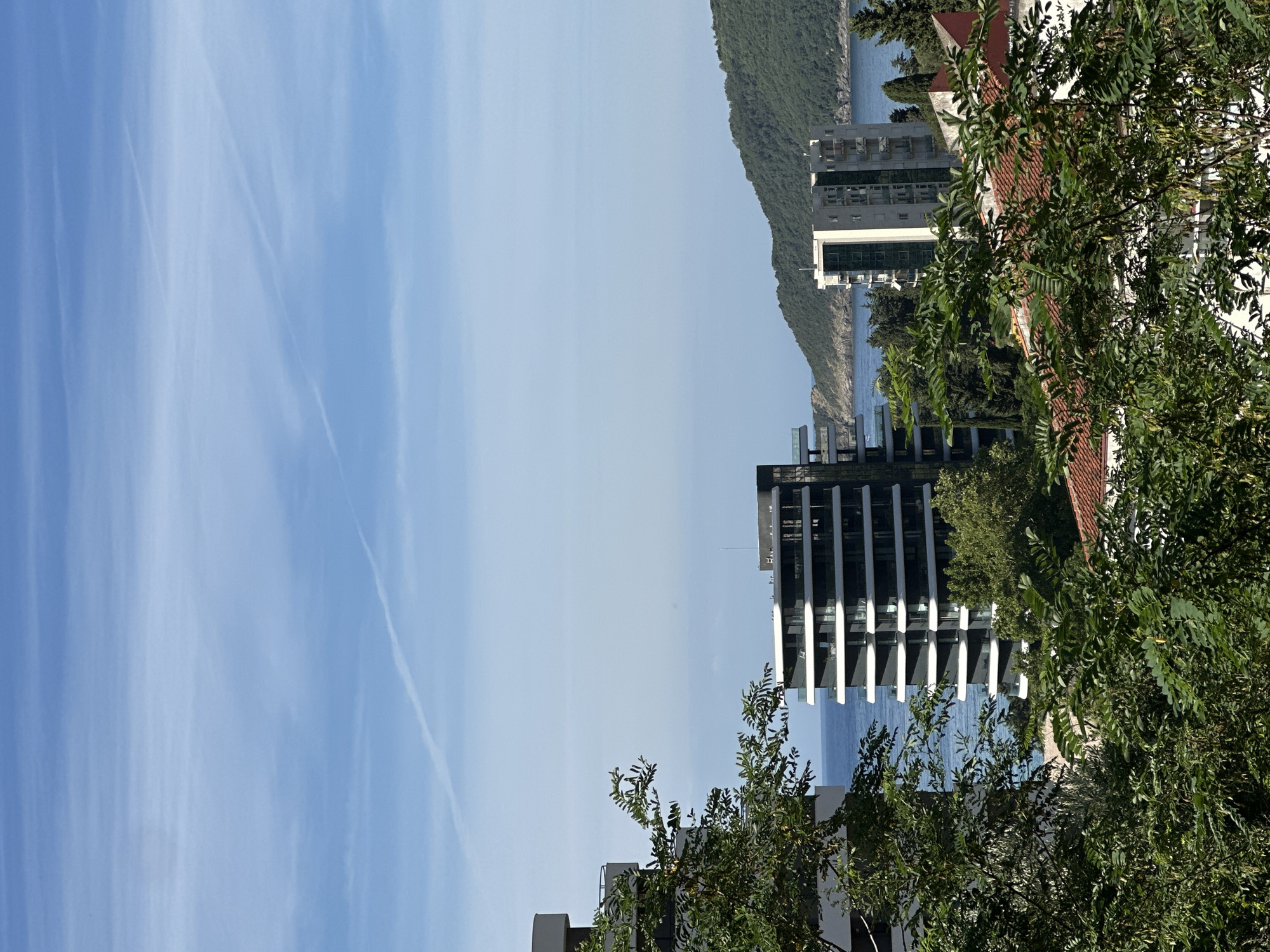 The image captures a scenic view of Budva, Montenegro, focusing on a modern apartment complex and its surroundings. In the foreground, dense green foliage of trees partially obscures the view, with some leaves and branches clearly visible and in sharp focus. Behind the foliage, a contemporary apartment building with a black facade and white balconies stands prominently. To its right, another apartment building, lighter in color, with visible balconies and glass windows, is situated.
Beyond the buildings, the calm, blue sea stretches towards the horizon, meeting a clear, light blue sky. The sky is marked with thin, white contrails from aircraft, indicating a bright, sunny day. A verdant, tree-covered hill rises from the sea in the mid-ground, adding a natural element to the urban landscape. The architecture suggests a residential area, possibly with a view of the coastline. The overall impression is one of a modern coastal city blending with natural beauty.