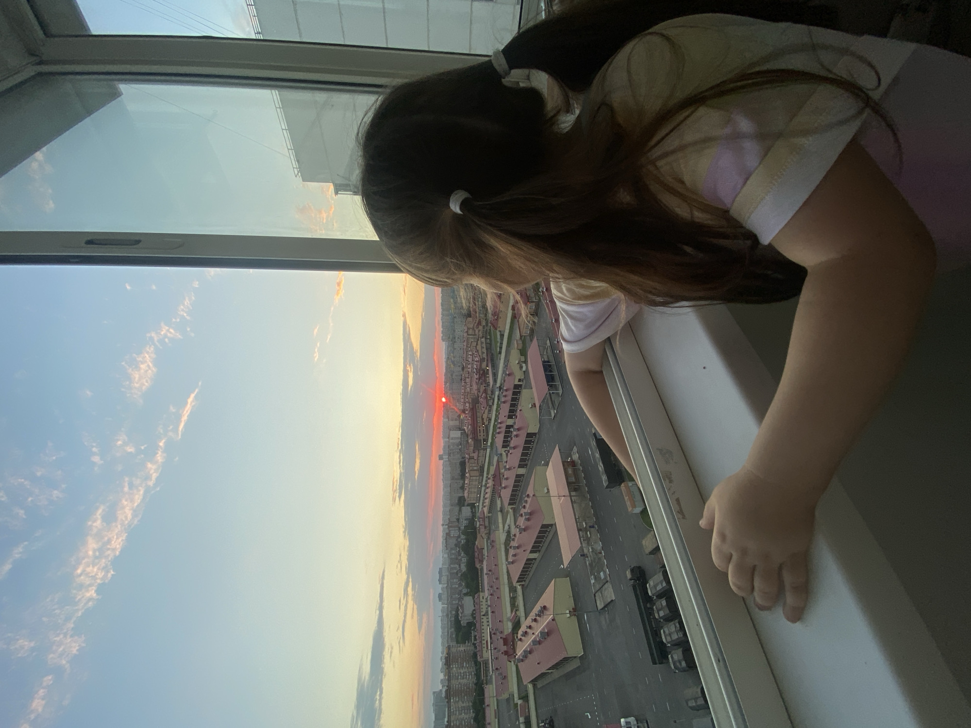 In the image, a young girl with long brown hair tied in two pigtails leans out of a window, looking down at the cityscape below. She is wearing a t-shirt with horizontal stripes of pastel colors. The sun appears to be setting, casting a warm glow on the sky and the buildings. The city below consists of numerous multi-story buildings, some with red roofs, arranged in a grid-like pattern. A paved road with parked vehicles is visible between the buildings. The sky is a blend of soft blues and oranges, with wispy clouds. The girl's pose suggests curiosity and perhaps wonder as she observes the view. The setting is an urban environment, likely a residential area, viewed from a high vantage point, characteristic of an apartment building in Krasnodar, Russia. The time of day is clearly sunset, indicated by the colors in the sky and the low angle of the sun, which is visible as a bright red orb on the horizon.