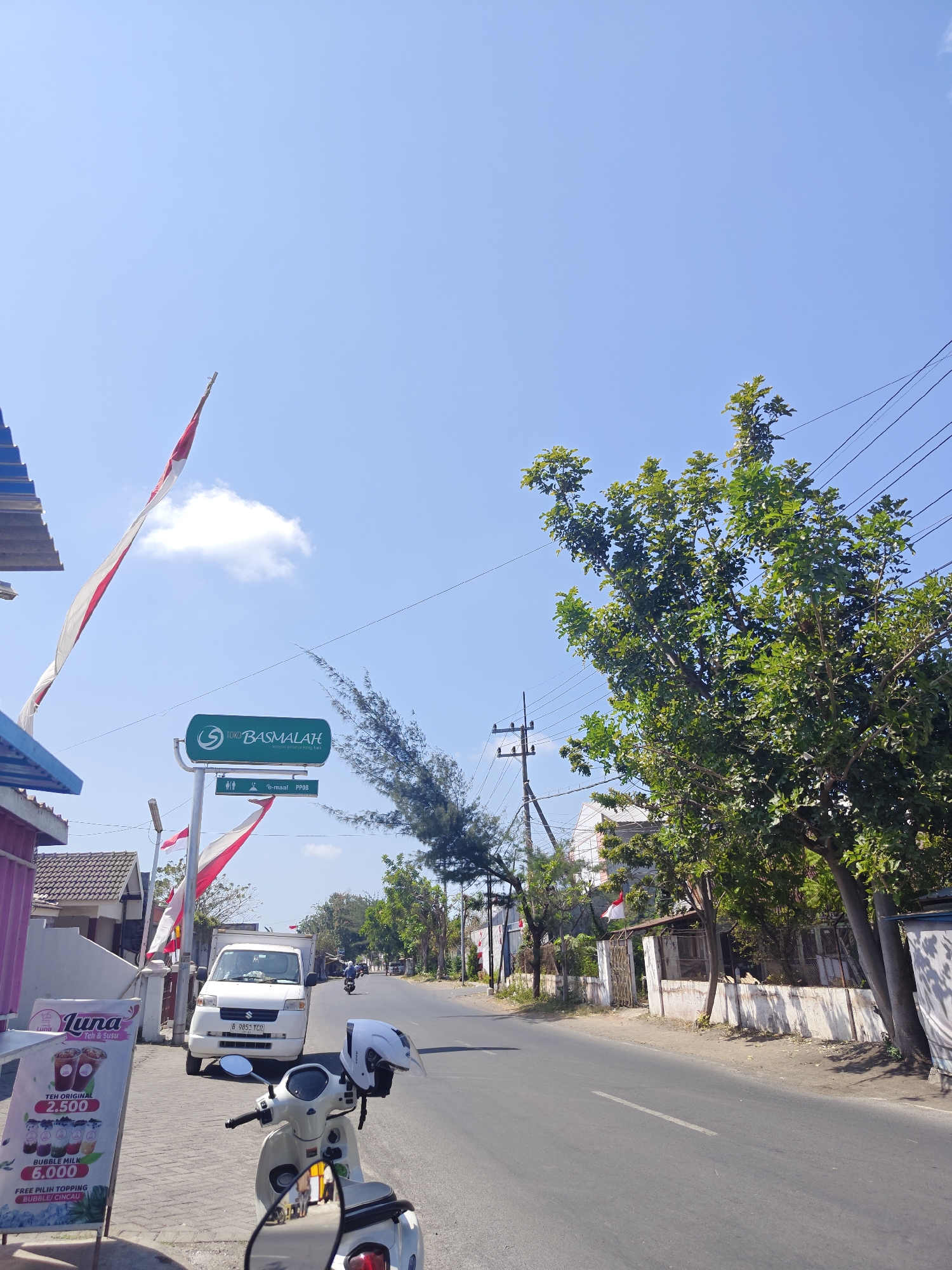 The image shows a street scene in Situbondo, Indonesia, on a bright, clear day. On the left side of the street, there is a storefront with a sign that reads "Luna Teh & Susu" advertising drinks with prices. Next to it, a white scooter with a white helmet on the handlebars is parked. Further down the sidewalk, a white delivery van is parked. A tall green sign with "TOKO BASMALAH" and smaller icons indicates a business. Two Indonesian flags, red and white, are flying from poles, one large one on the left and a smaller one near the sign.
The street itself is paved and appears to be a two-lane road with a white dashed line marking the center. The right side of the street is lined with trees, some of which are full of green leaves, and a white-walled building with a fence. Power lines are visible running overhead. In the distance, traffic can be seen, including a motorcycle with a rider. The overall environment is a sunny, roadside setting in a town or city.