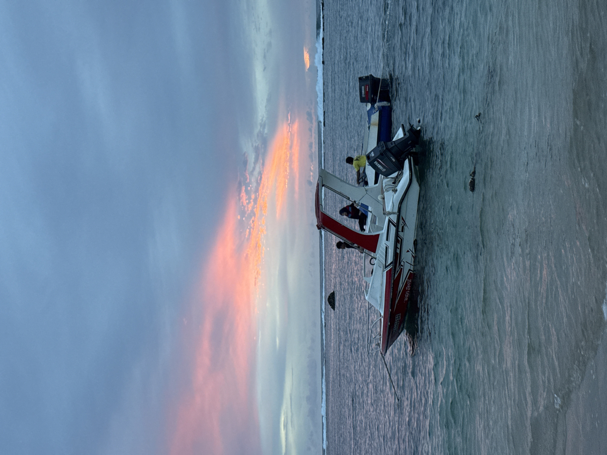 The image depicts a serene ocean scene at dusk, with two boats anchored in the calm water. The sky is a canvas of soft blues and pinks, with streaks of orange suggesting the setting sun. In the foreground, a white and red speed boat is partially visible, with a person seated inside. Behind it, a blue and white boat has a person in a yellow shirt standing. The water is a deep blue, with gentle waves lapping against the shore in the distance. The overall atmosphere is tranquil and picturesque, evoking a sense of peace and relaxation. The image does not provide specific details or context that would allow for the identification of the location as Meunasahbale, Indonesia.