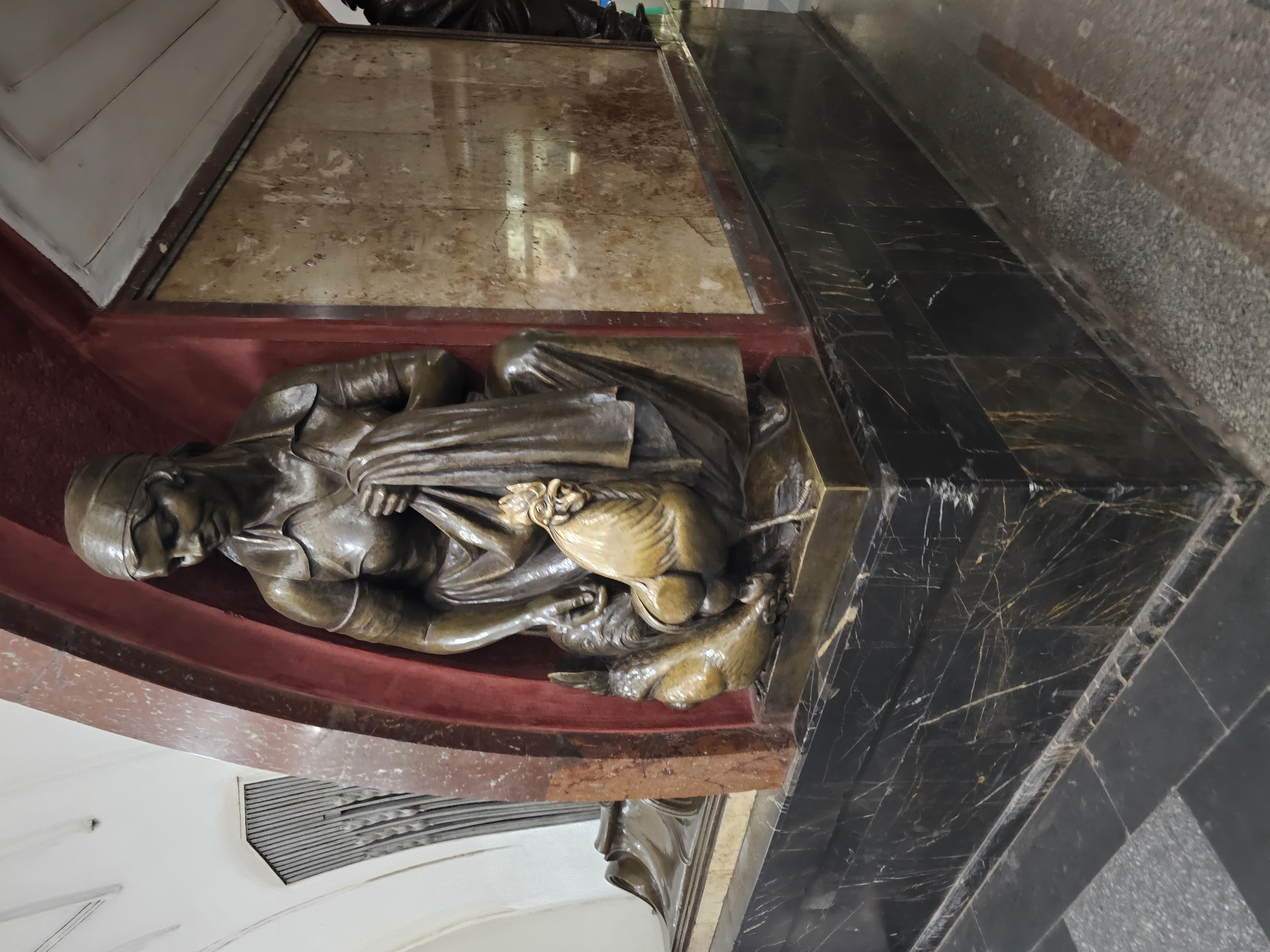 A bronze sculpture depicts a woman kneeling, holding a piece of fabric. She is accompanied by two chickens, one large and one small. The sculpture is situated in an alcove with red and marble accents. The base of the sculpture is made of black marble with gold veining. The background includes a marble wall and a tiled floor. The scene is indoors, suggesting a public building or a museum.
