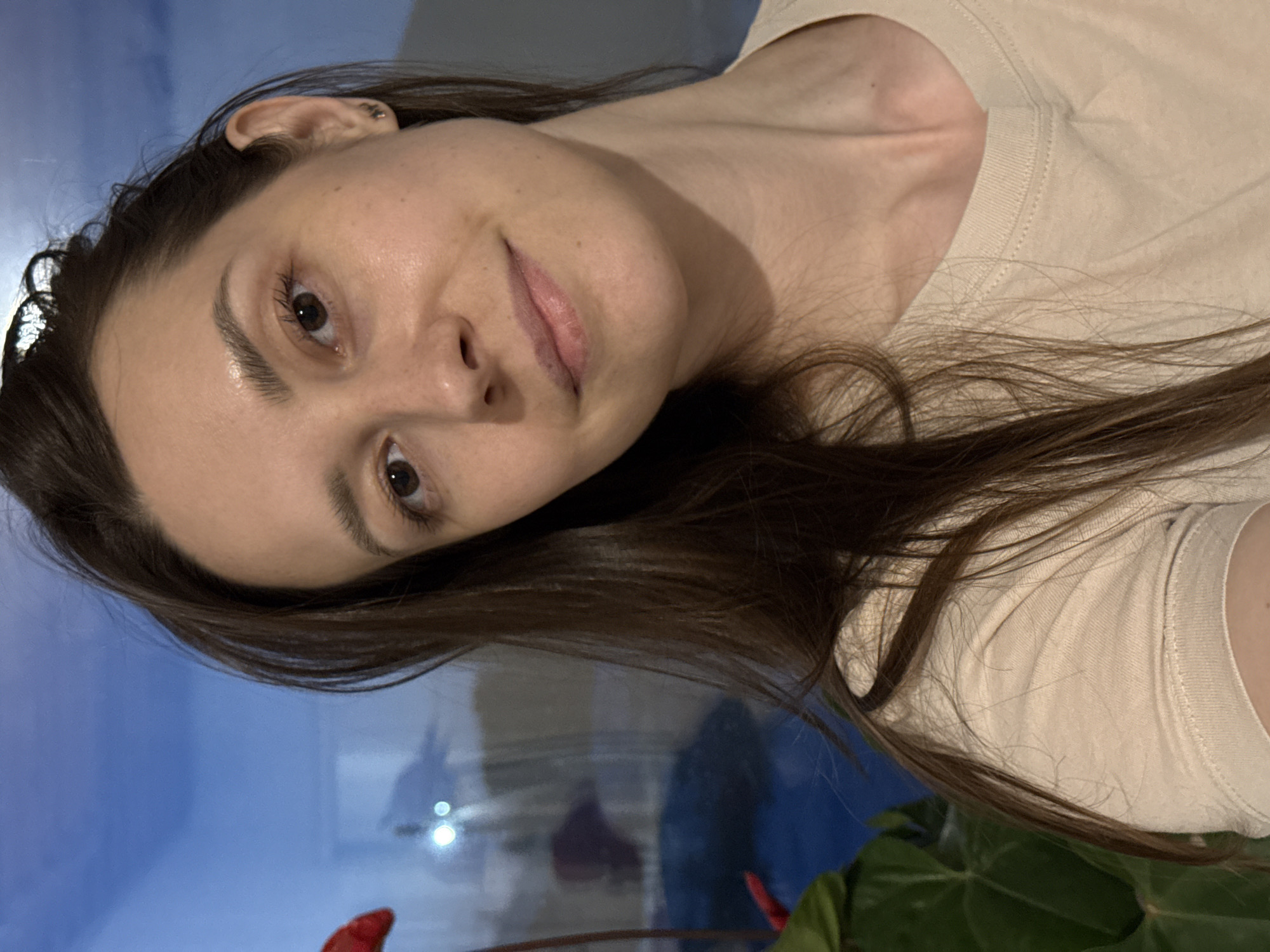 A woman with brown hair and eyes is shown in a selfie. She is wearing a light-colored t-shirt. The background appears to be a window with a blue sky and a reflection of a building. Part of a red flower and green leaves are visible at the bottom. No specific details indicate the location is Chaikovsky, Russia.