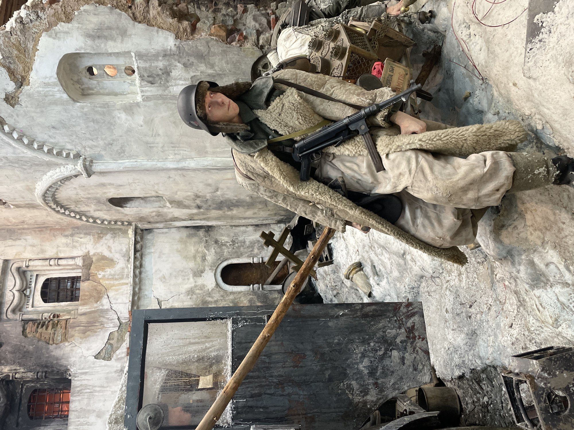 The image depicts a diorama of a World War II scene, likely located in Moscow, Russia, given the presence of a Soviet soldier. The soldier is dressed in a winter uniform, including a fur-lined coat and helmet, and is armed with a submachine gun, likely a PPSh-41 or similar. The background shows a war-torn building, possibly a church or other historically significant structure, with damaged walls and windows. The scene suggests urban combat, with debris and wreckage visible. The presence of religious symbols like a cross further indicates the setting as a place of worship or cultural importance. The overall tone is somber and reflective of the intense battles fought during the war.