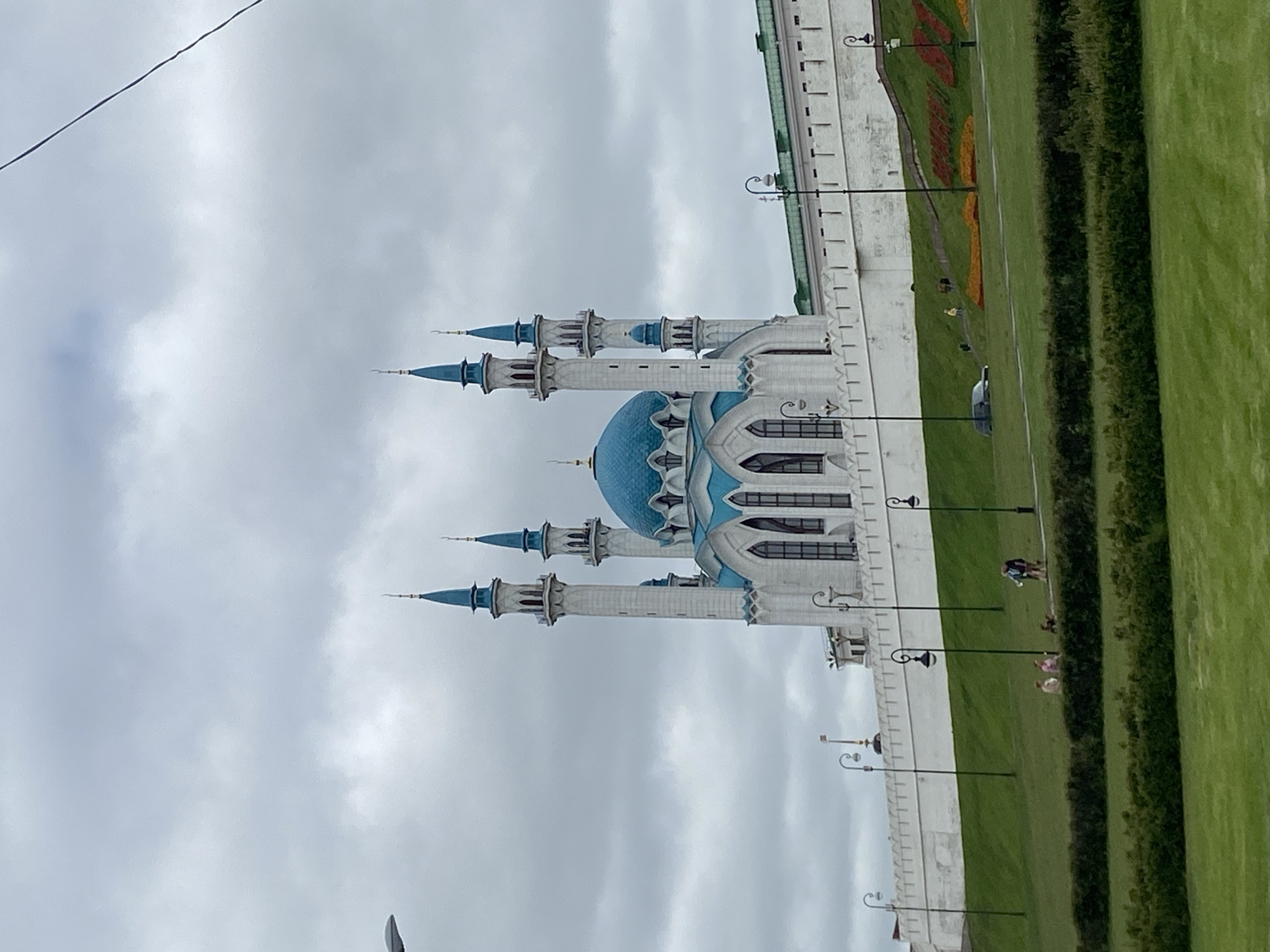 The image, when correctly oriented, presents a scenic view of a prominent architectural structure, likely the Kul Sharif Mosque, situated in Kazan, Russia. The mosque is a striking focal point, characterized by its elegant light-colored facade, a distinctive blue main dome, and multiple slender minarets topped with matching blue spires. Ornate detailing is visible on the mosque's architecture and windows.
To the right of the mosque, a sturdy white or light grey wall extends horizontally, indicating a larger complex or fortress, consistent with the Kazan Kremlin setting. Along the base of this wall and extending into the foreground are expansive, meticulously maintained green lawns. Vibrant flower beds arranged in patterns of red, orange, and yellow add splashes of color to the landscape, contrasting with the greenery. Neatly trimmed hedges form distinct borders within the lawn area.
Several streetlights with traditional, curved designs are placed at regular intervals along the wall and pathways. A few individuals are casually present on the green lawn; one person in dark clothing is visible near the flower beds, while others, possibly two figures, can be seen further down near the hedges.
The sky is overcast with a consistent light grey cloud cover, suggesting a cloudy day. A thin, dark power line or wire diagonally crosses the upper left portion of the sky. The overall atmosphere appears peaceful and serene, showcasing a well-preserved public space surrounding a significant landmark. No visible text is present in the image.