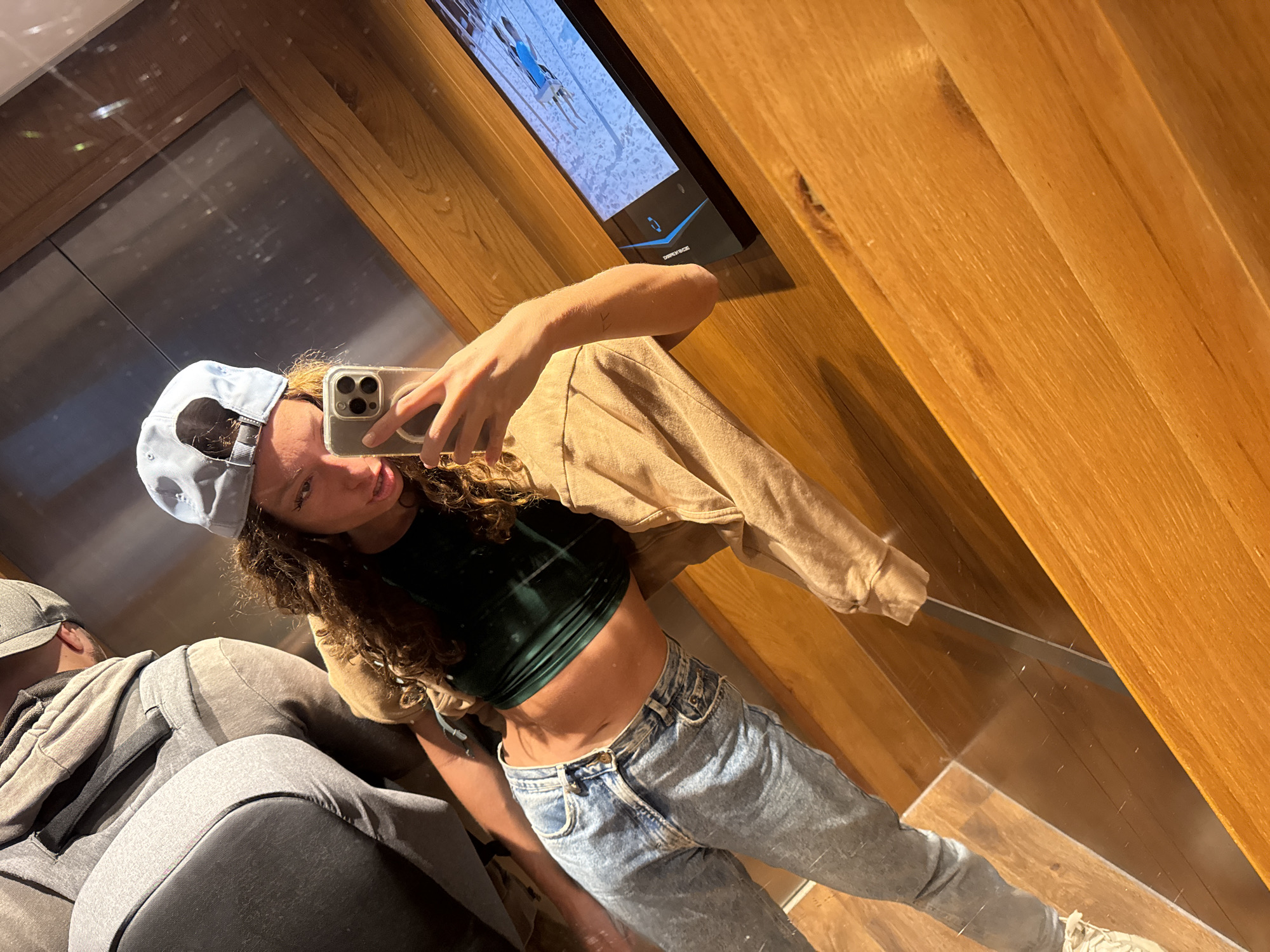 A young woman is taking a selfie in an elevator. She is wearing a light blue baseball cap, a dark green crop top, and light blue jeans. She is holding a smartphone in her right hand. She has long, curly brown hair. The elevator has wooden paneling on one side and a mirror on the other. There is a screen mounted on the wall of the elevator displaying a video. There is another person seated behind her in the elevator. The location is not definitively identifiable as Đà Nẵng, Vietnam from the image alone.