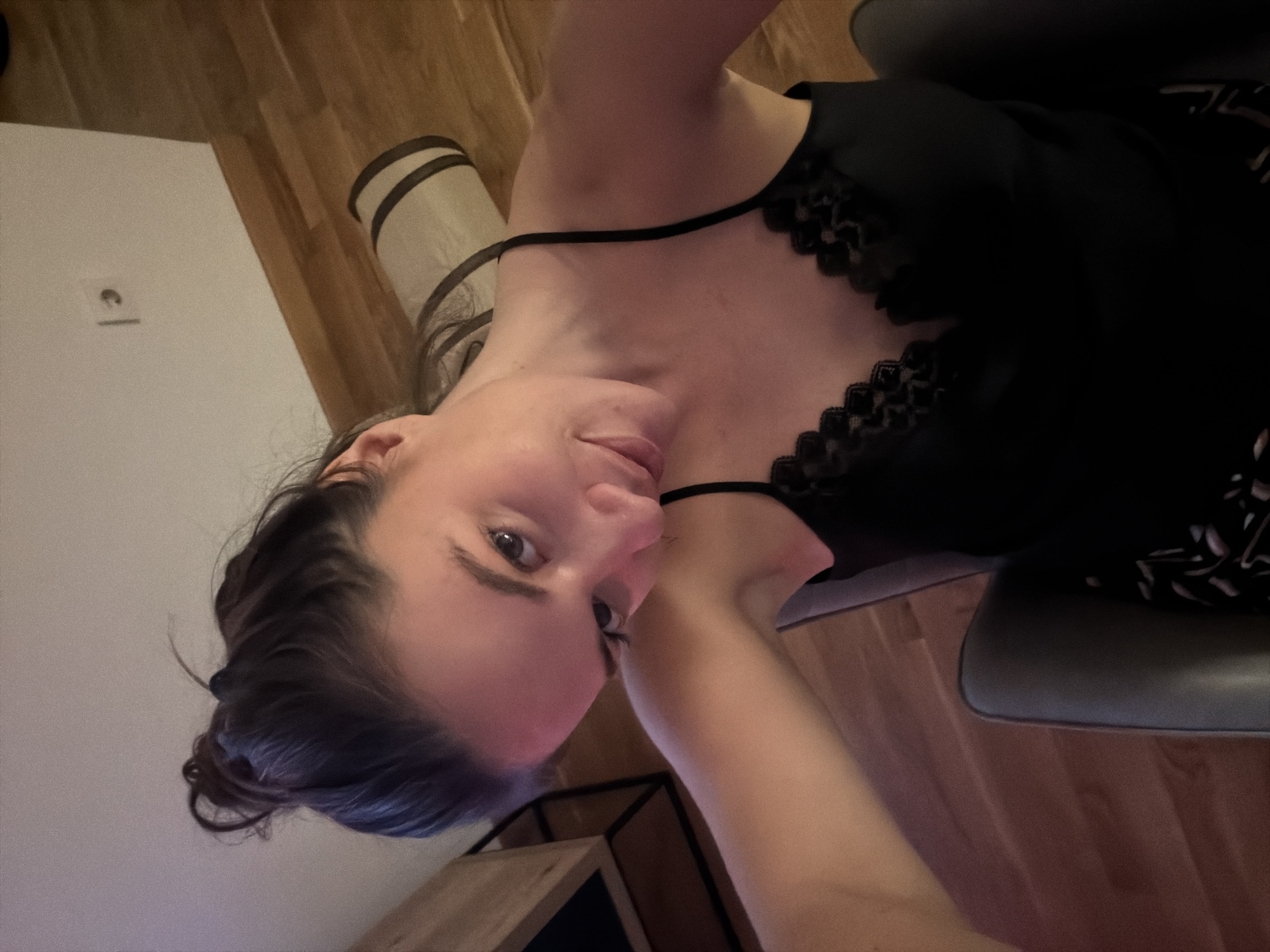 A woman is seated indoors. She is fair-skinned, with dark hair pulled back in a bun and is wearing a black camisole with lace trim. The background reveals a wooden floor and a white wall with an electrical outlet. The scene appears to be indoors, possibly in an apartment or home. The location in the city of Belgrade, Serbia cannot be determined based on the visual information provided.
