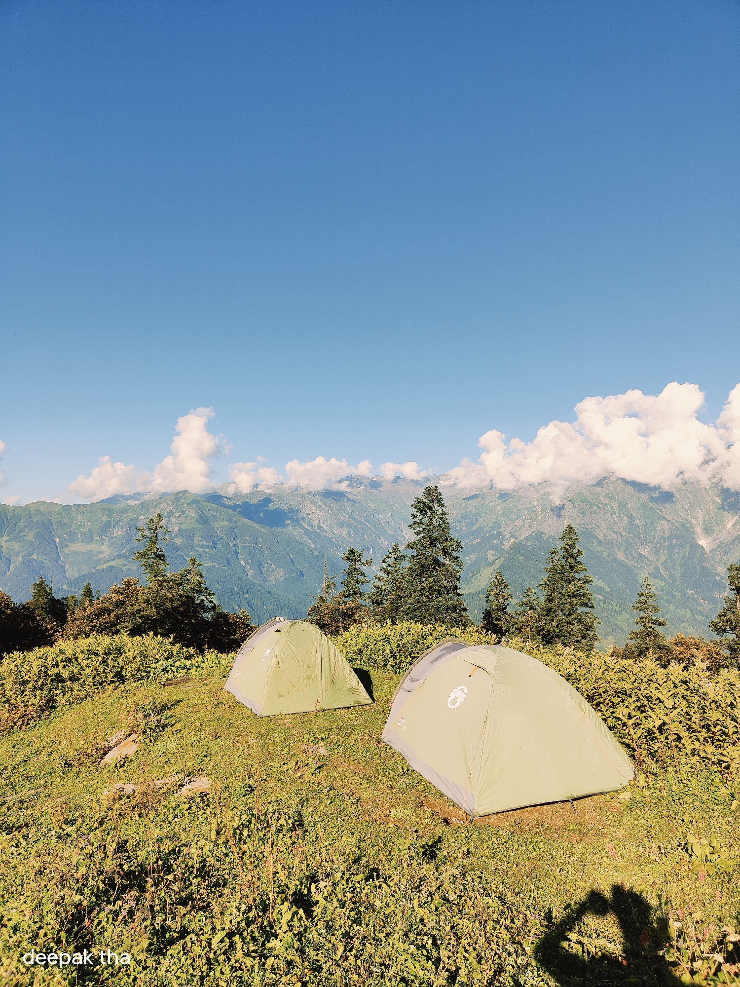 Two light green tents are pitched on a grassy slope. The tents are facing slightly towards the viewer, with the tent on the right being closer and larger than the one on the left. The tent on the right displays a circular logo. The ground is covered in lush green grass and small flowering plants, with some rocks visible on the slope. In the background, a dense forest of pine trees stands before a majestic mountain range. The mountains are covered in greenery and some patches of snow near the peaks, with white clouds clinging to them. The sky above is a clear, bright blue with a few scattered white clouds. The bottom left corner of the image has a watermark that reads "deepak tha". A shadow of a person, possibly holding a camera, is visible in the bottom right foreground, creating a heart-like shape. The scene appears to be a campsite in a mountainous region, likely near Manali, India, given the context provided.