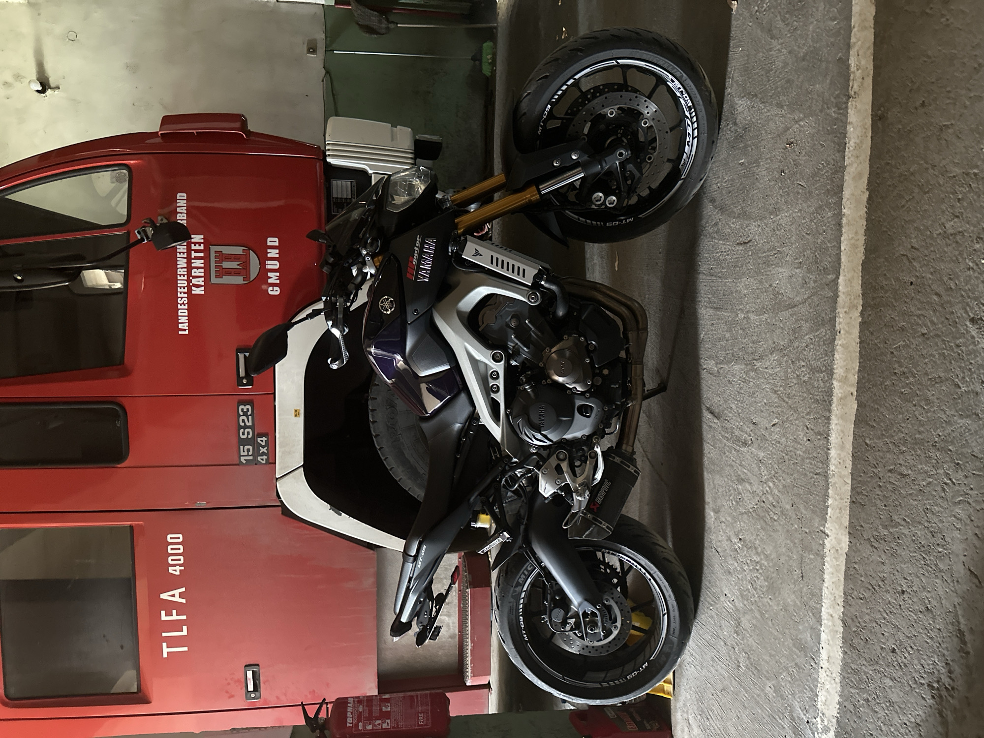 A red fire truck is parked behind a dark purple and silver Yamaha motorcycle in what appears to be a garage or workshop. The fire truck has text on its side including "TLFA 4000", "15 S23 4X4", "LANDESFEUERWEHRBAND KÄRNTEN", and "GMÜND". The motorcycle has "YAMAHA" and "MT-09" logos visible. A fire extinguisher is mounted on a stand to the left of the motorcycle, and a broom is leaning against the wall to the right. The floor is concrete, with a white line painted across it. The overall lighting suggests an indoor setting. The image does not depict the city of Valchedram, Bulgaria.